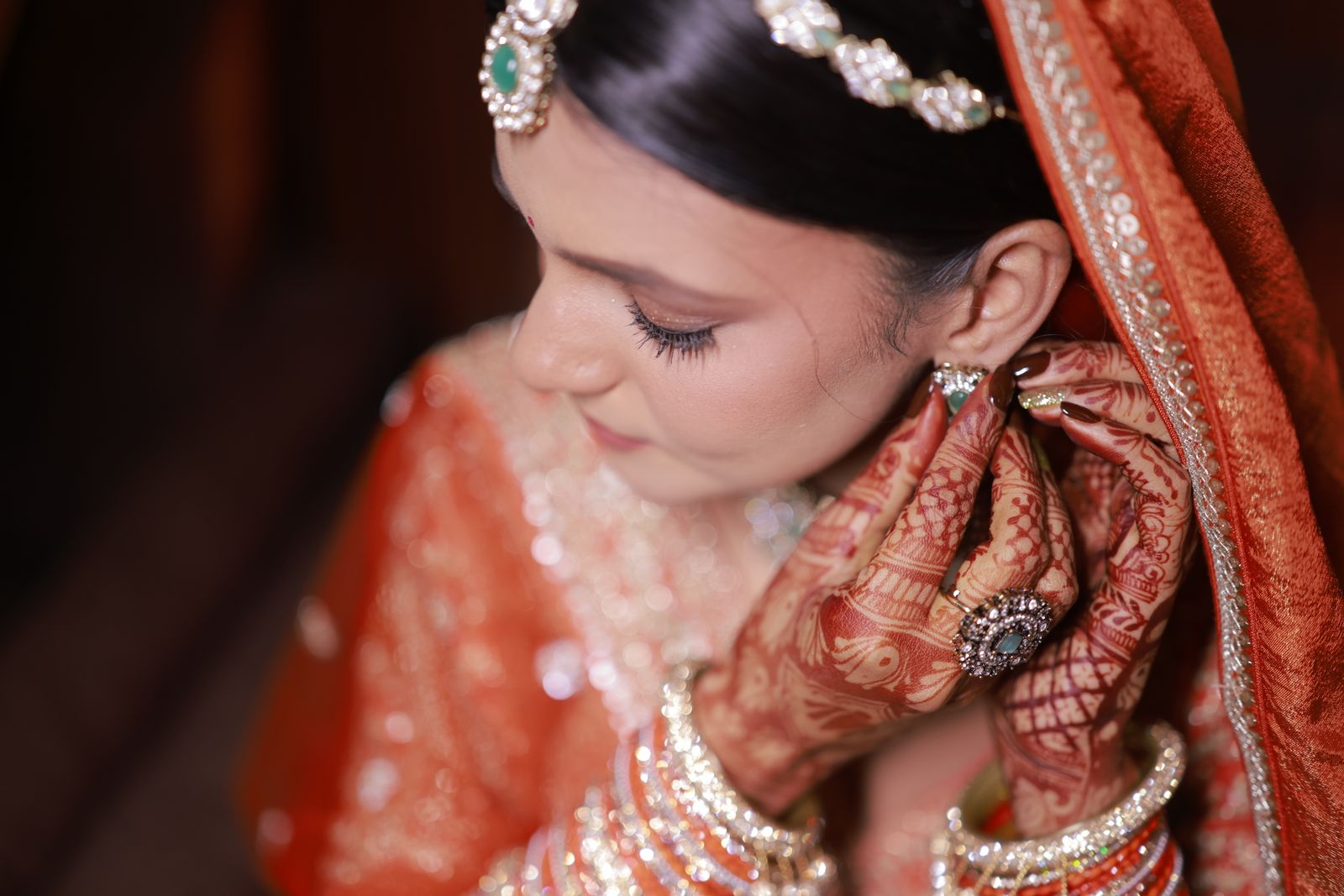 A bride in a burnt orange dupatta fastens an earring, both hands covered in mehndi, the room falling away dark behind her.
