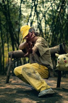 A young adult wears casual attire, poses outdoors in a bamboo forest beside a plush toy, embodying playful spirit.