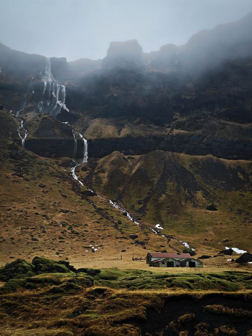Free A lone cabin sits beneath majestic mist-covered mountains with cascading waterfalls. Stock Photo