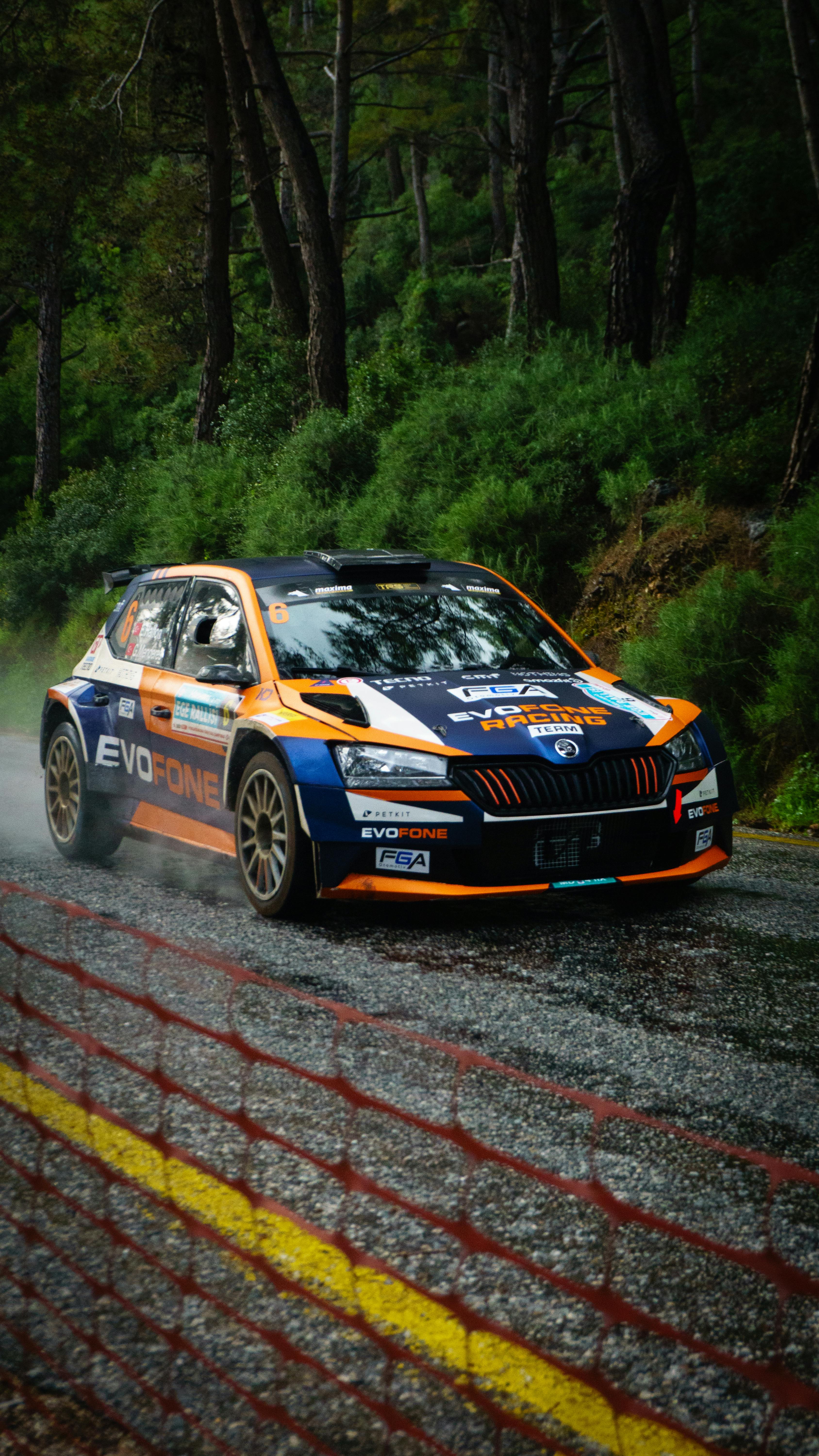 A vibrant rally car driving through a forest on a wet road, showcasing thrilling motorsport action.