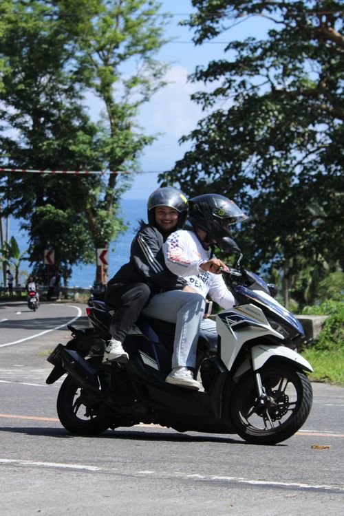 Free Two adults riding a motorcycle along a scenic coastal road under bright sunlight. Stock Photo