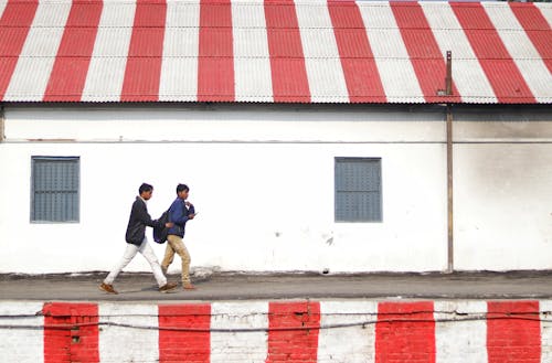 Free Side view of young Asian guys walking on platform painted with white and red stripes along shabby stone building with striped white and red roof on daytime Stock Photo