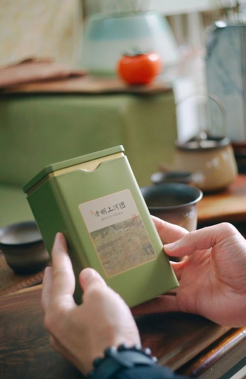 Free A person holds a green tea tin in a cozy, tea-drinking scene with a teapot and cups in the background. Stock Photo