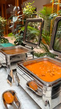 Elegant buffet display with stainless steel chafing dishes featuring curry dishes, surrounded by lush greenery.