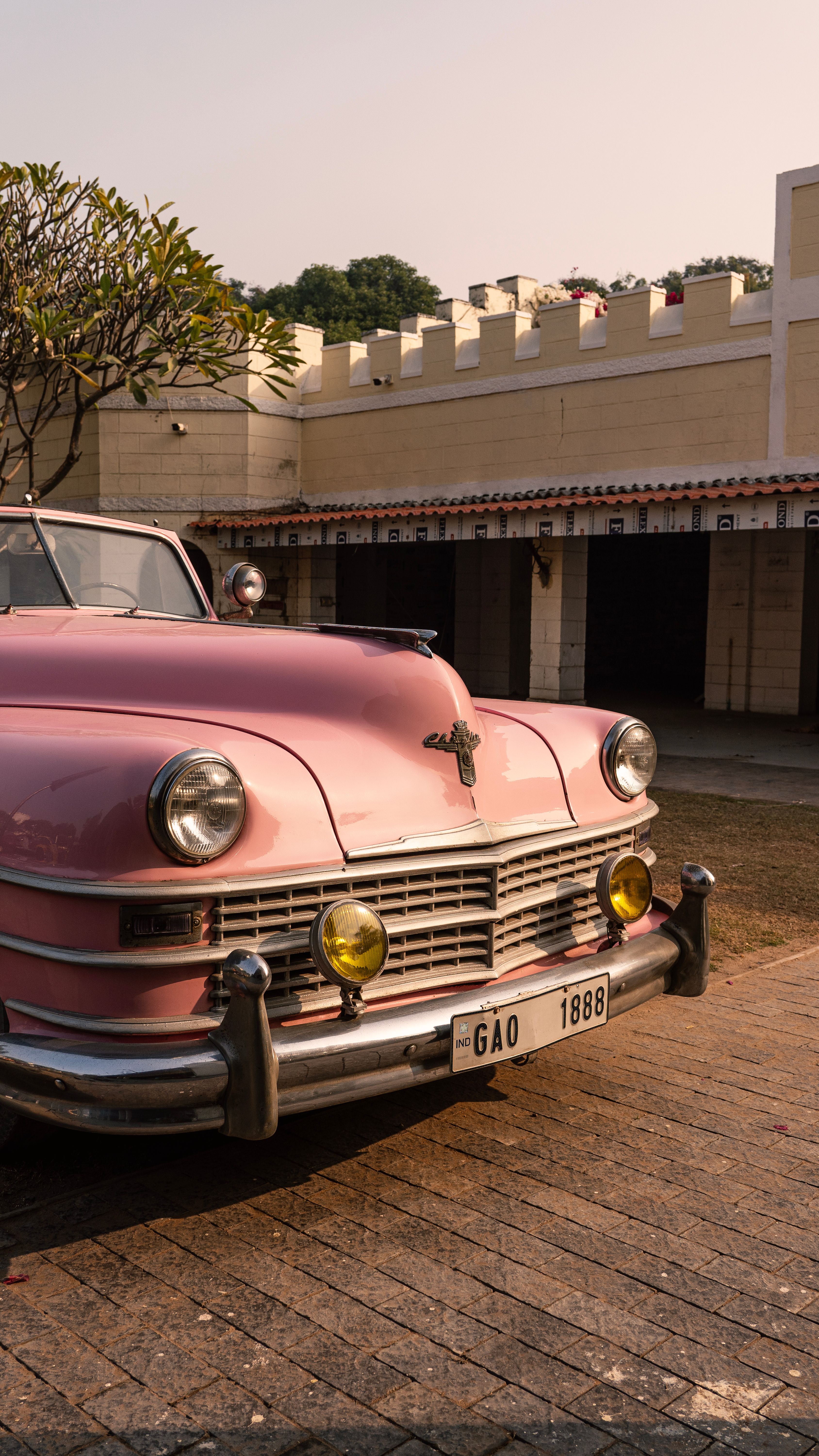Charming pink vintage car parked at a heritage location in India, with warm sunlight and architectural backdrop.
