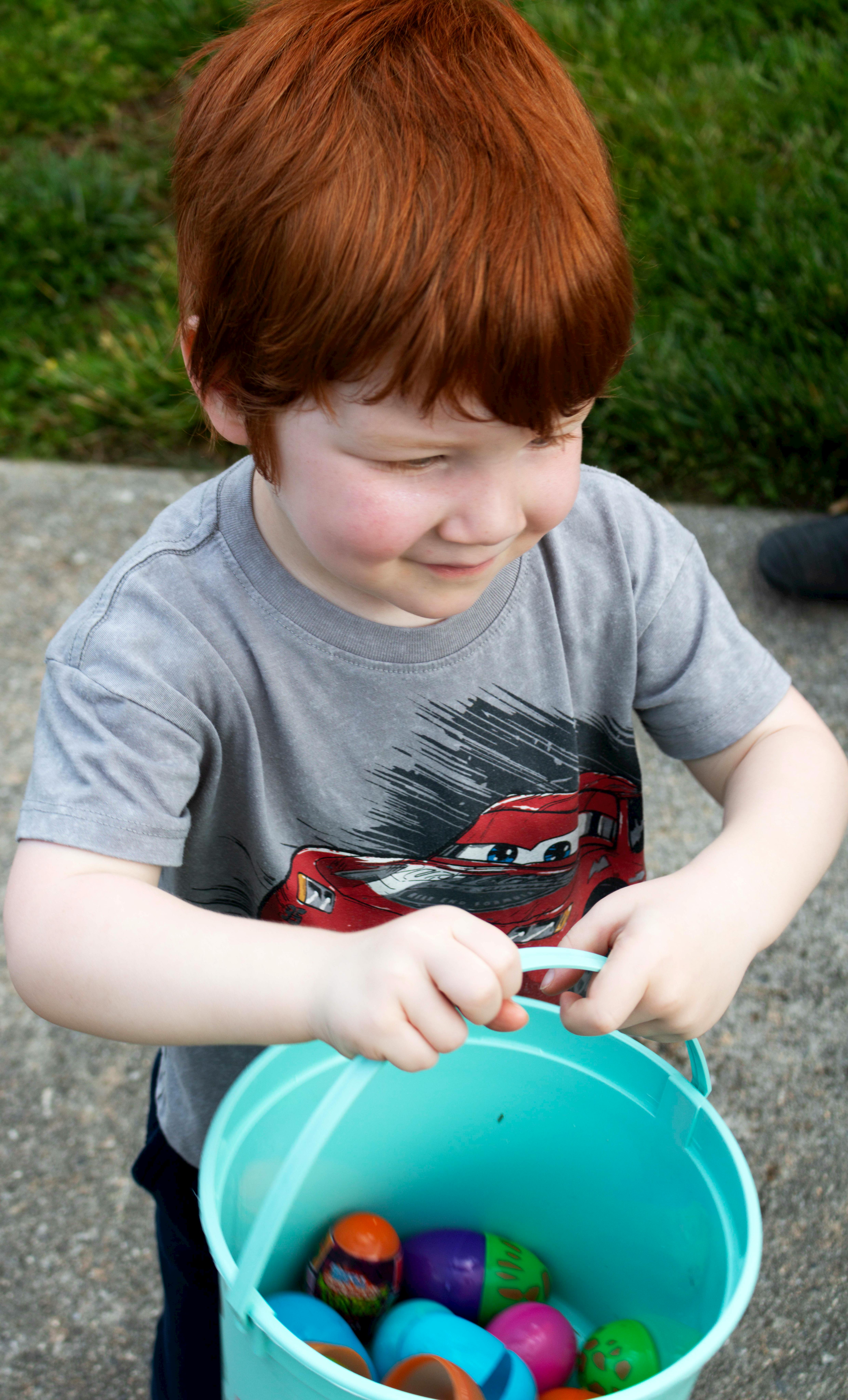 Young child with red hair collecting colorful Easter eggs in a blue bucket outdoors.