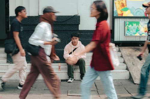 Free A young man in a pink hoodie sitting on steps as people walk by in motion blur. Stock Photo