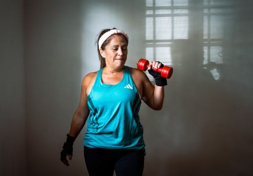 Middle-aged woman in workout attire lifting a red dumbbell. Indoor exercise scene promoting fitness and healthy lifestyle.