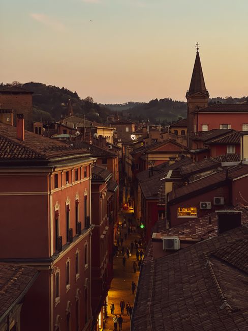 View of a historic street lined with buildings and a church spire at sunset in Bologna, Italy.