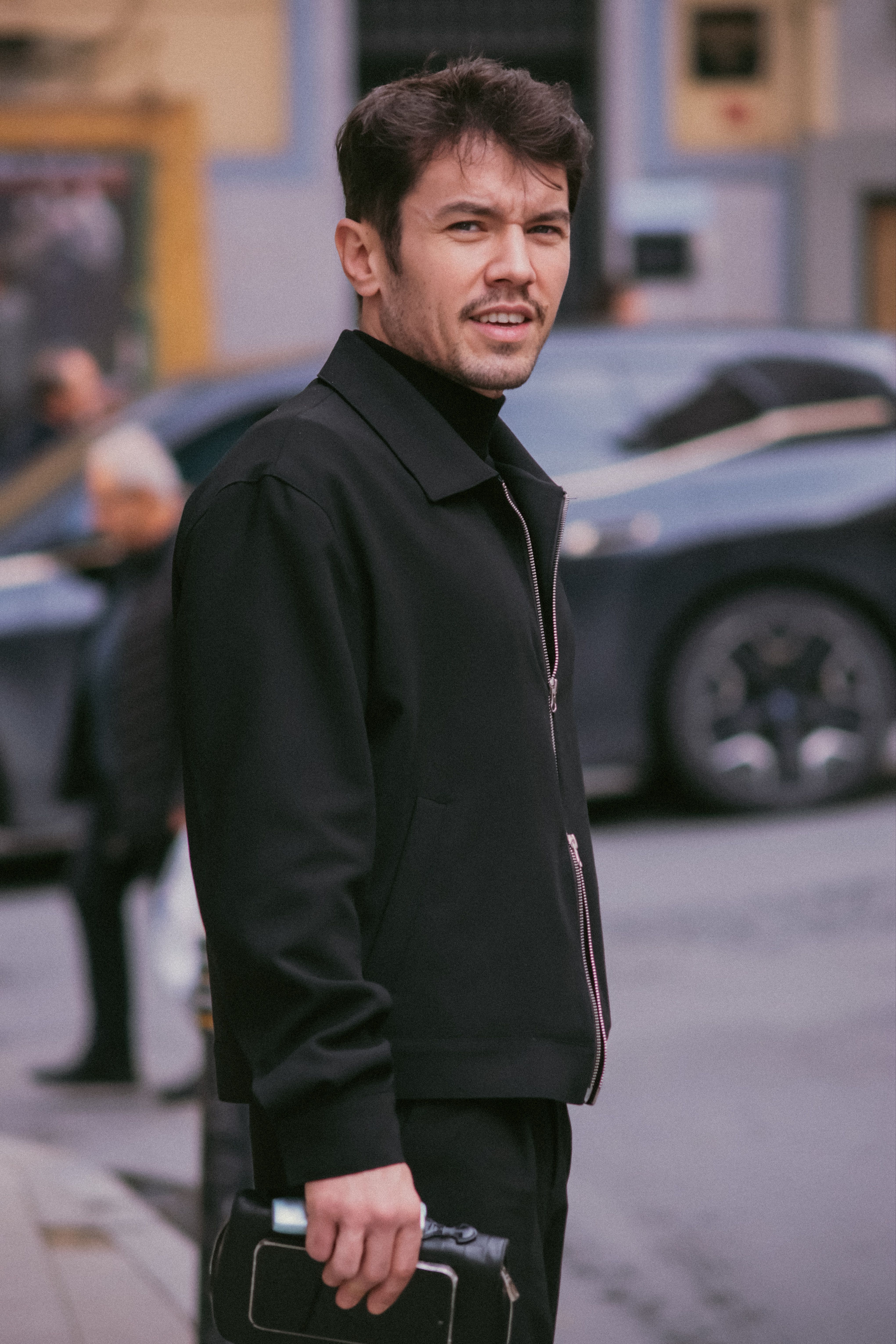 A man in a stylish black outfit stands on a busy urban street, holding a phone.