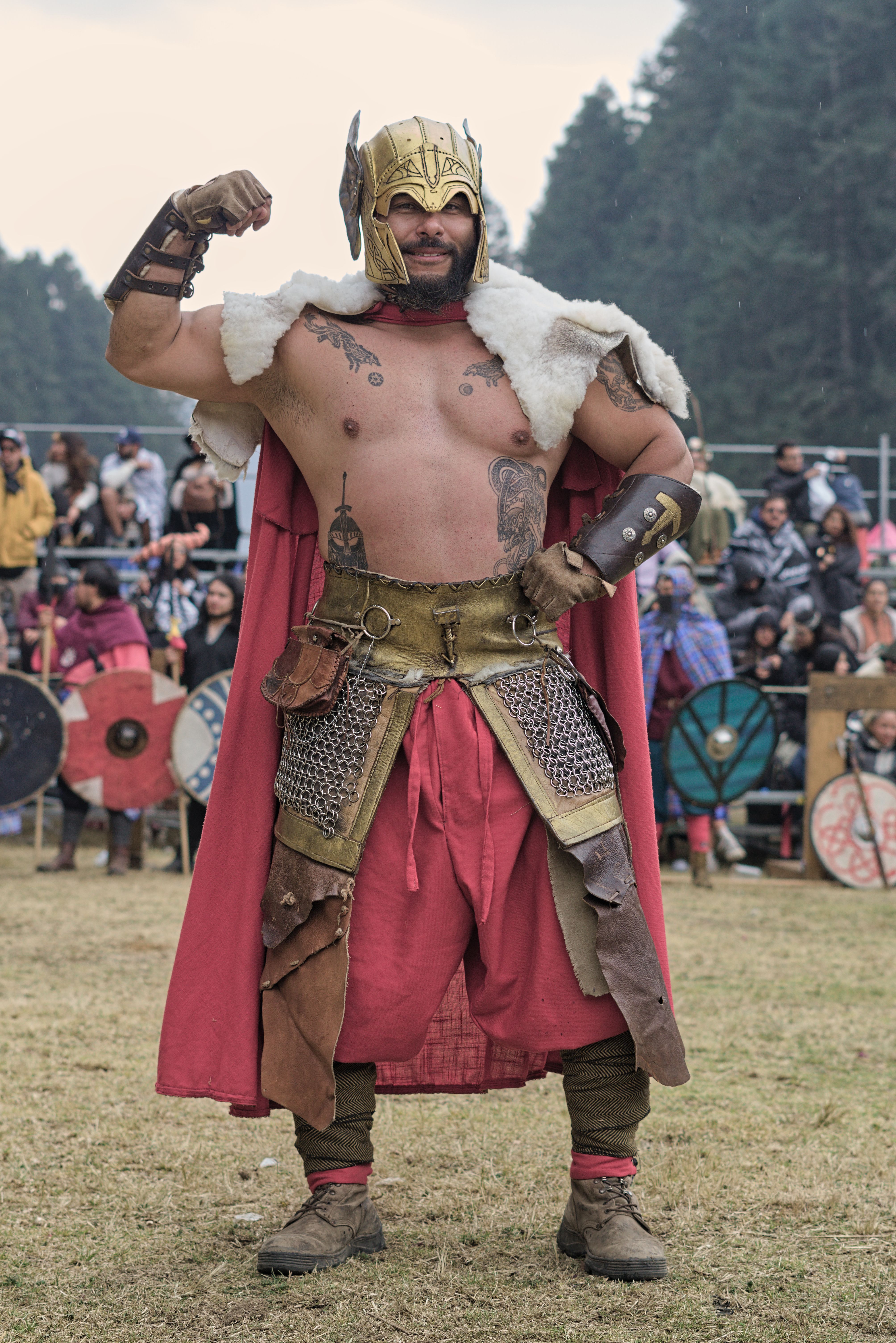A strong warrior in historical armor poses confidently with a crowd in the background at an outdoor festival.