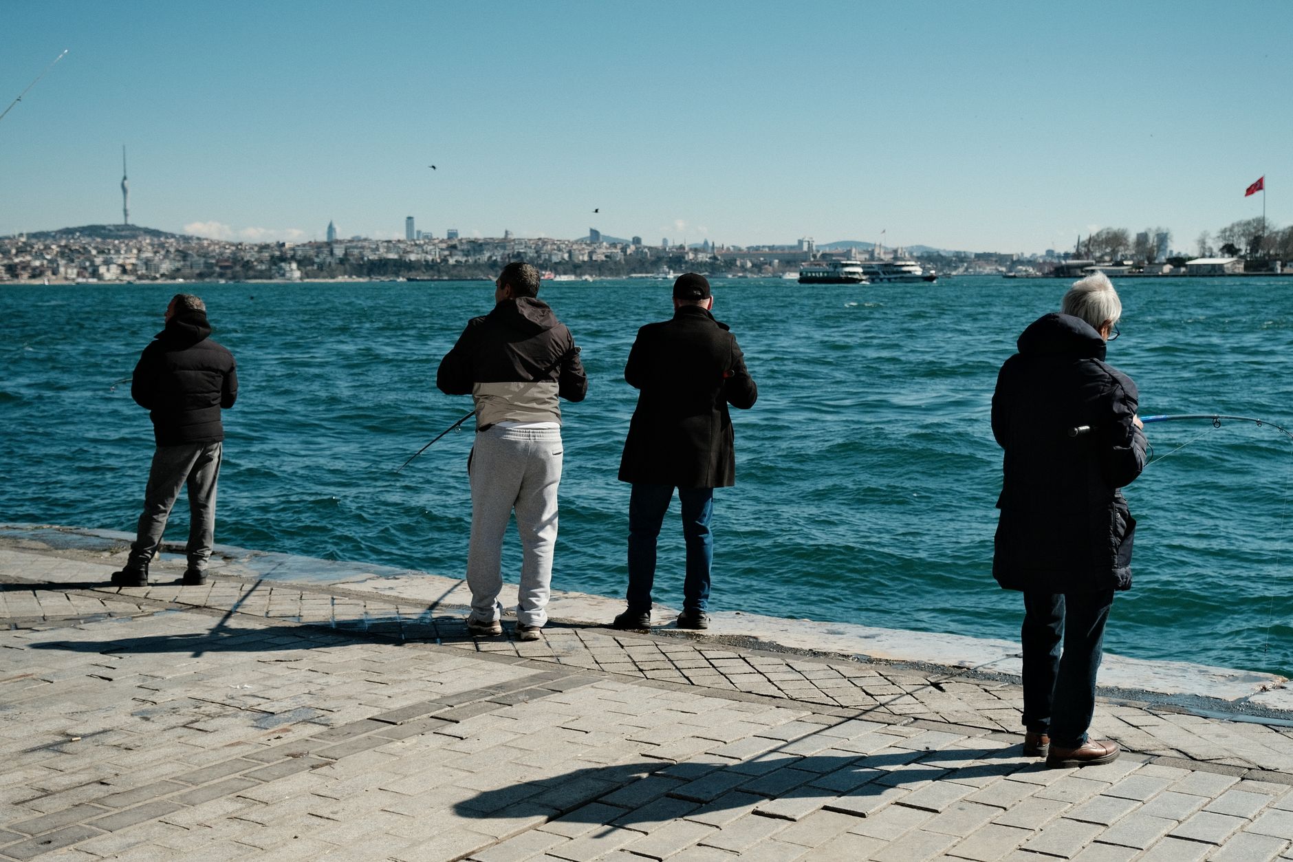 Four people fishing at the Bosphorus Strait with city skyline in the background on a sunny day.