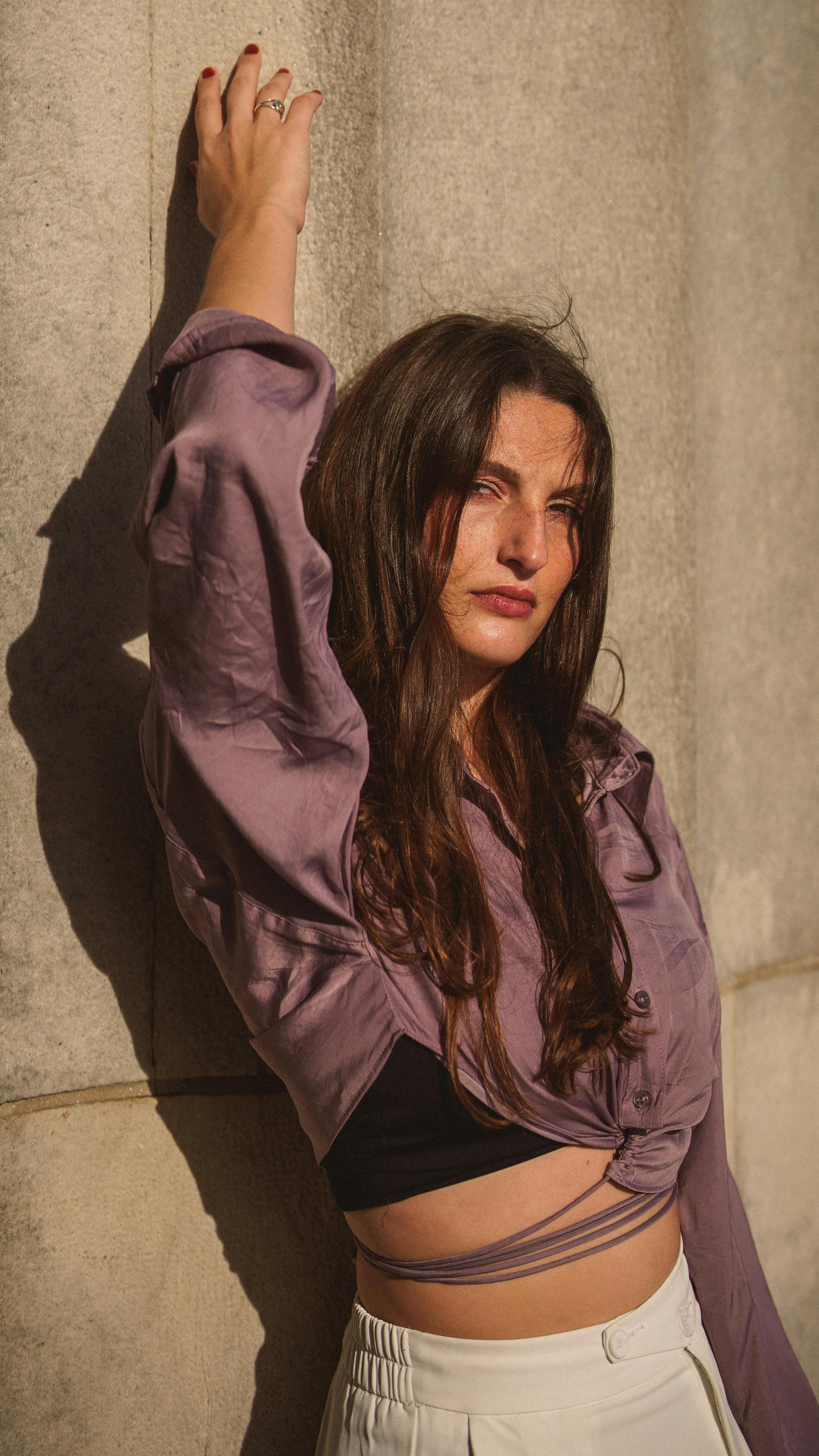 Stylish woman in purple blouse poses against stone wall in warm sunlight. Urban fashion.