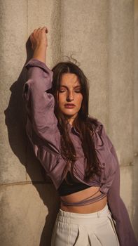 Fashionable woman in purple blouse poses against a stone wall in sunlight.