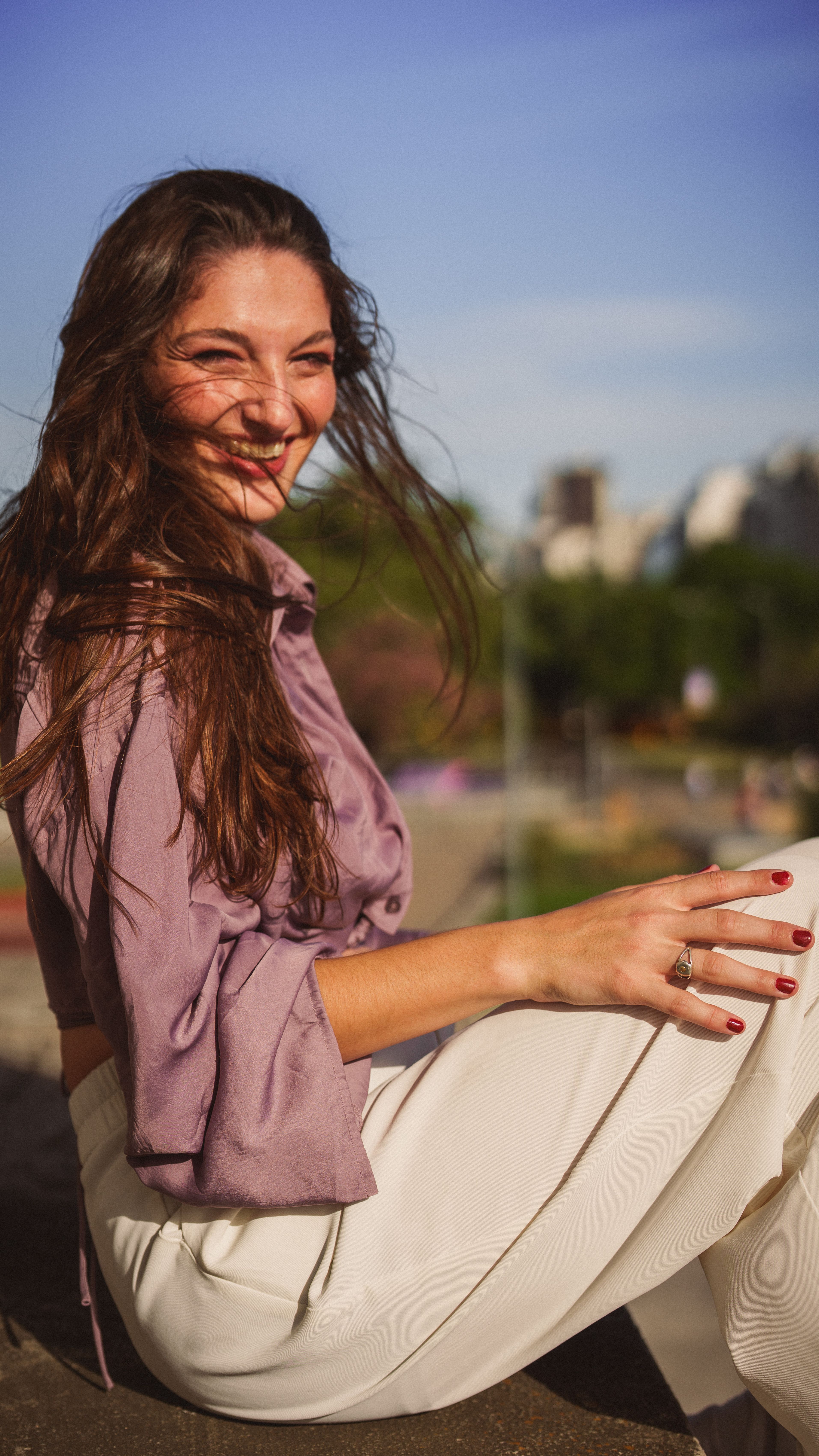 A joyful woman with a radiant smile sits in an urban park, basking in the sunlight.