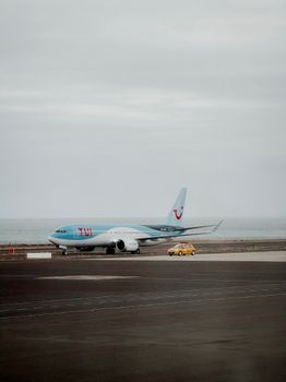 A commercial airplane and yellow vehicle on an airport runway with a scenic ocean backdrop and overcast sky.
