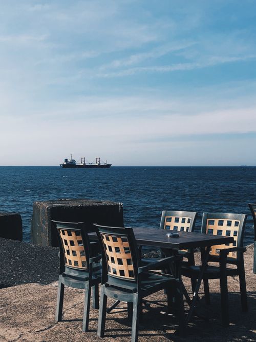 Free Seaside view with empty patio chairs overlooking a distant cargo ship under clear skies. Stock Photo