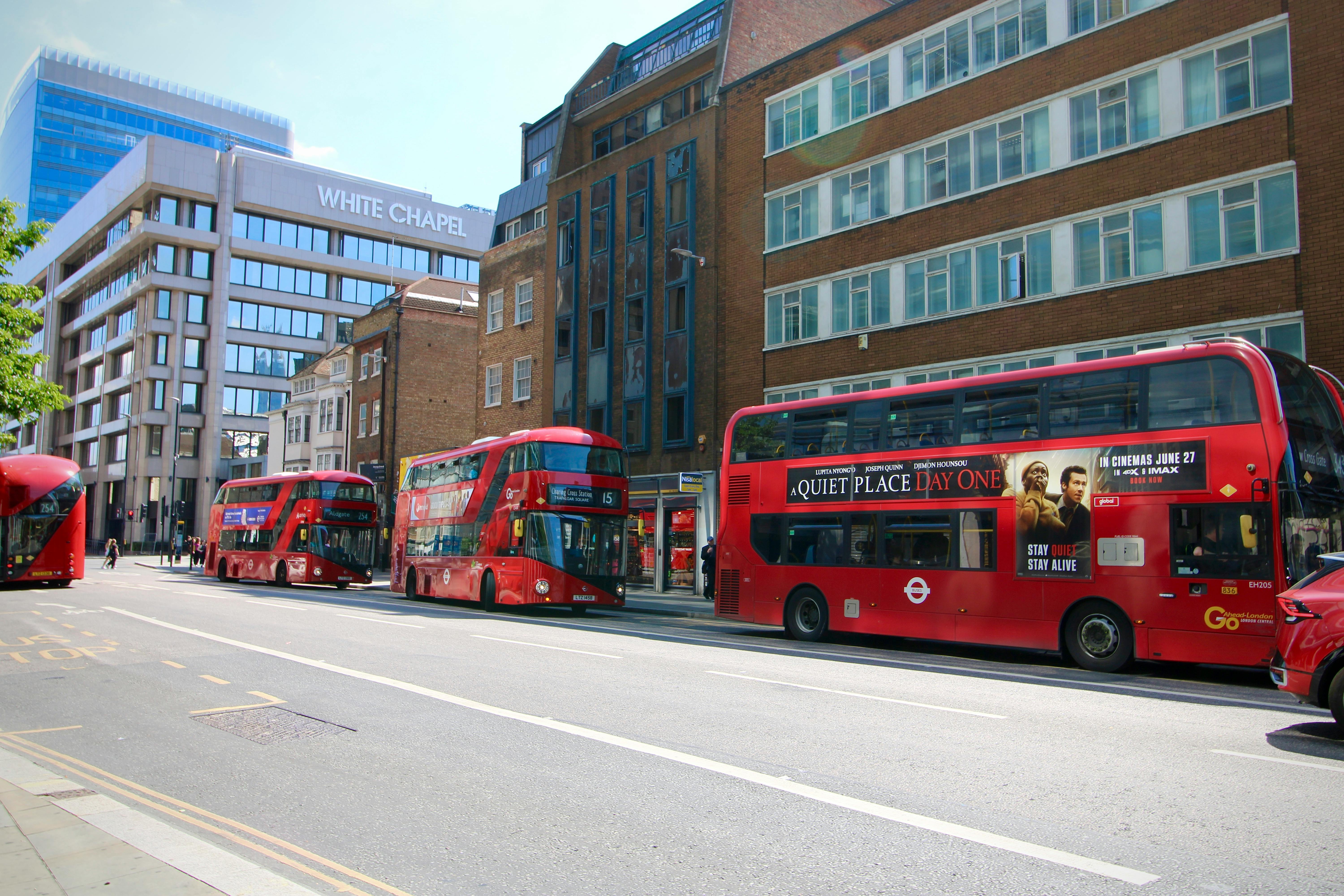 Red double-decker buses on a sunny day in Whitechapel, London, with city street and buildings.