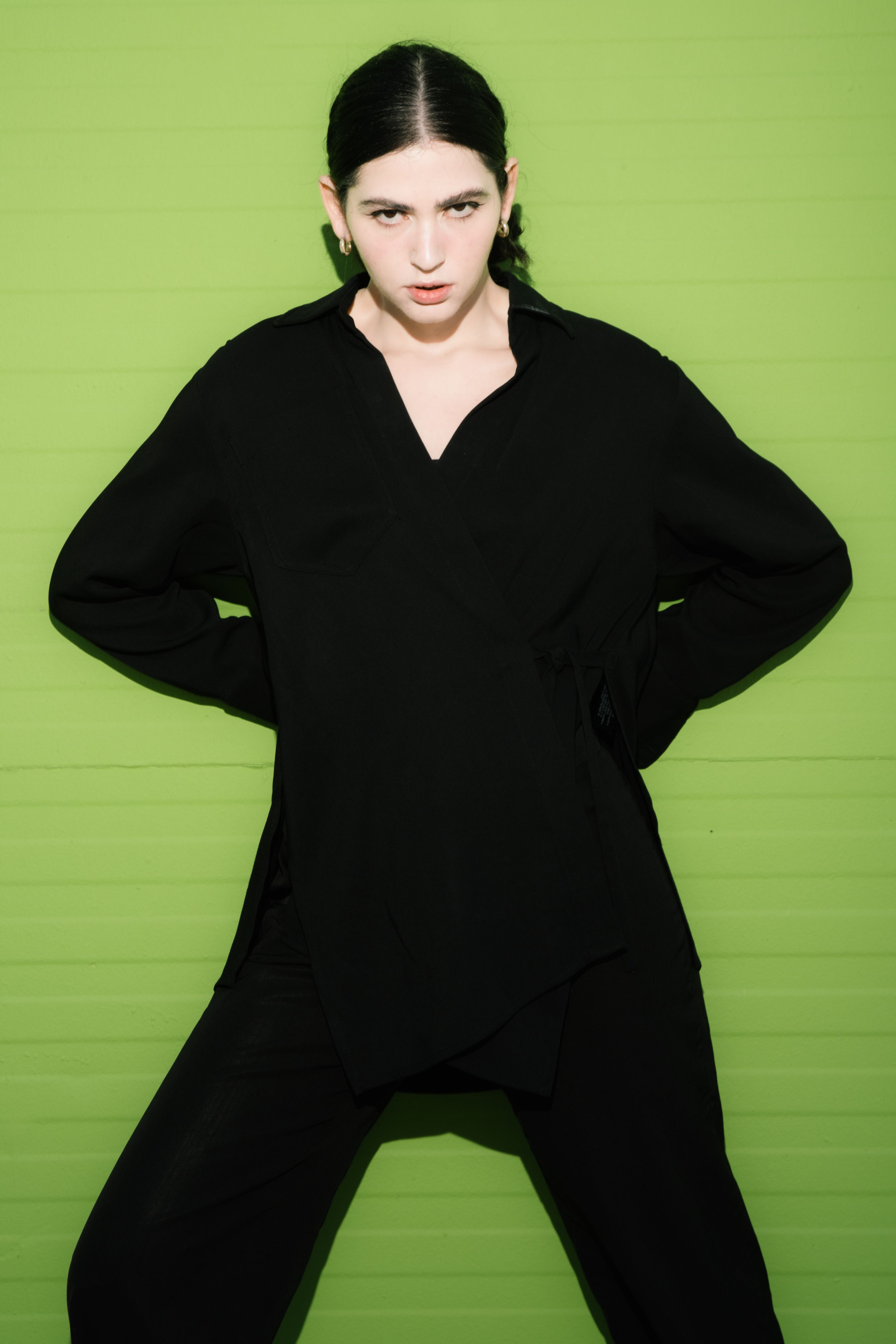 Stylish adult in black attire poses against green background, showcasing modern fashion.