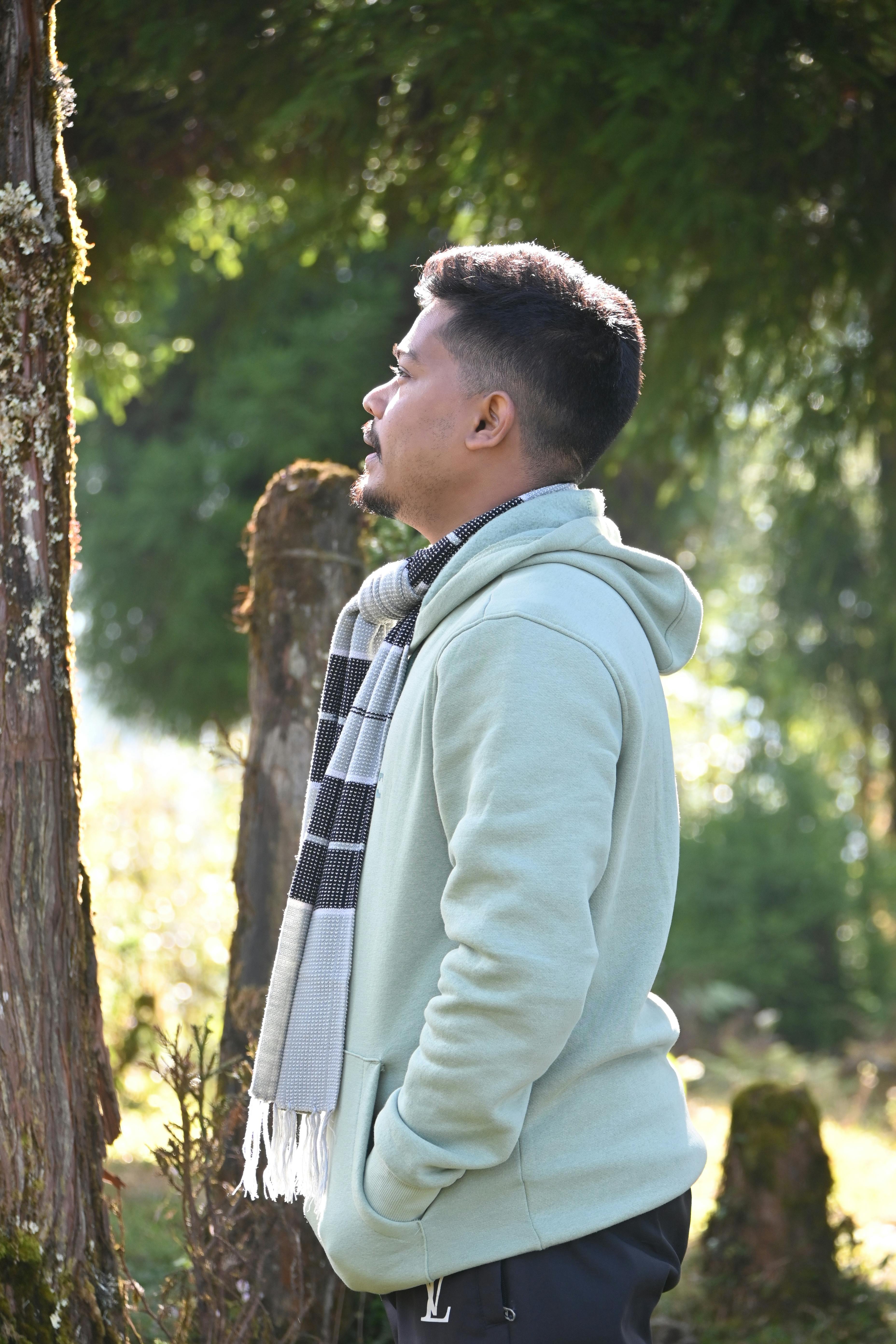 Profile view of a man in a light hoodie with a scarf, contemplating in a lush outdoor setting.
