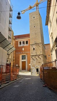 A narrow alley with construction barriers leading to an old stone tower in daylight.