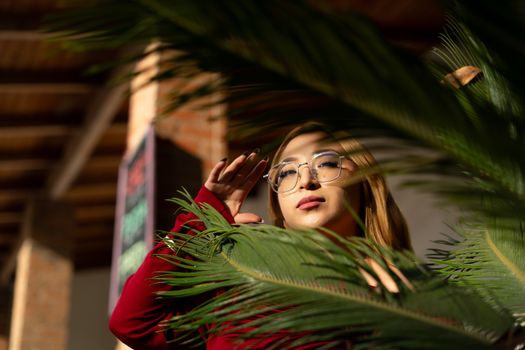 A stylish woman in glasses poses behind a lush fern indoors, illuminated by warm sunlight.