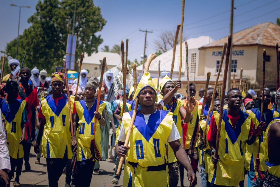 Vibrant cultural parade featuring participants in traditional yellow and blue attire holding wooden sticks.