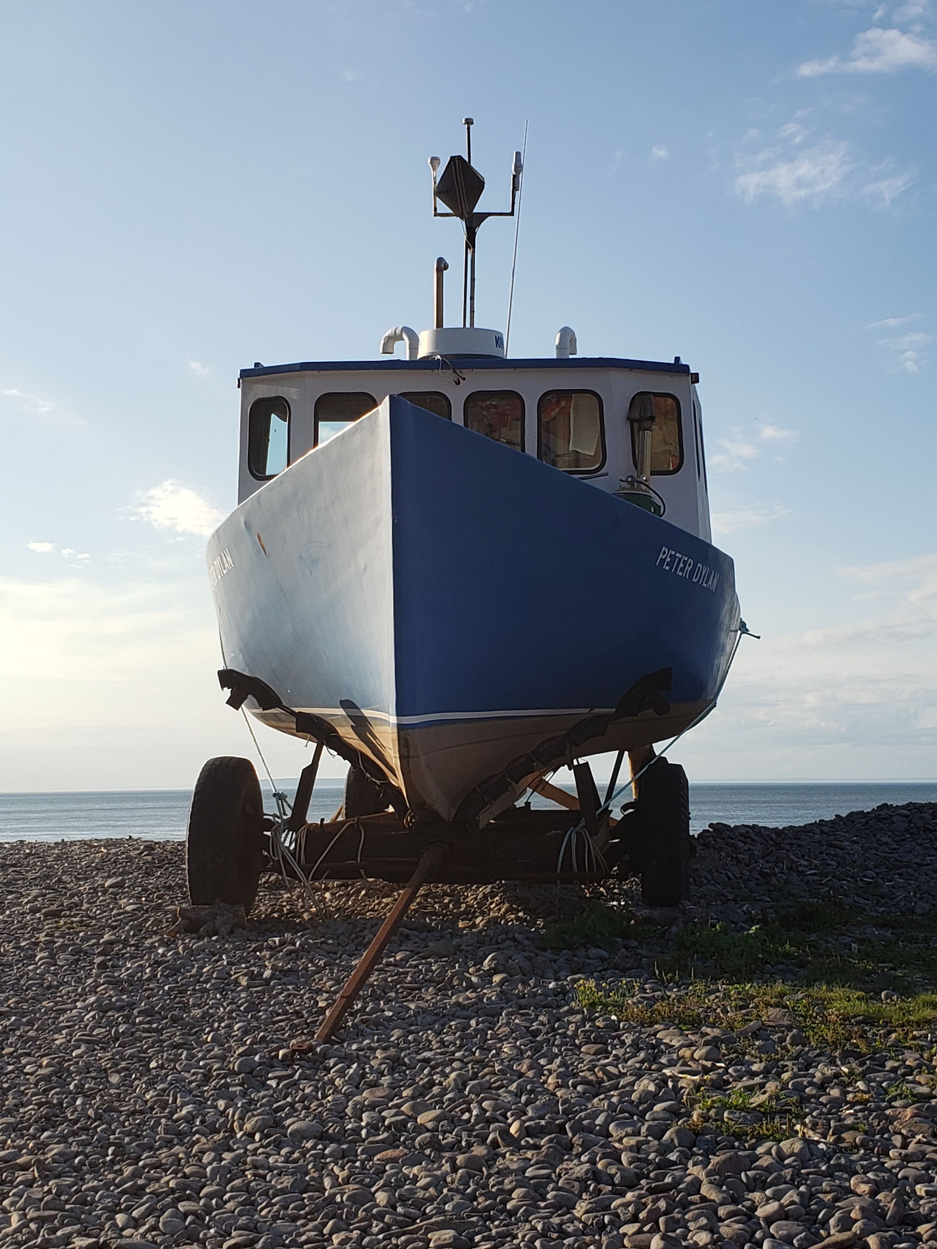 A blue fishing boat named 'Peter John' resting on a rocky beach against a serene sunset seascape.