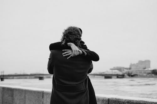 Grayscale Photo of Man Wearing Black Coat Hugging His Woman