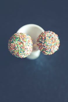 Two vibrant cake pops with colorful sprinkles displayed in a white cup, perfect for celebrations.