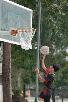 A teenager aims for a hoop in an outdoor basketball court with trees in the background.