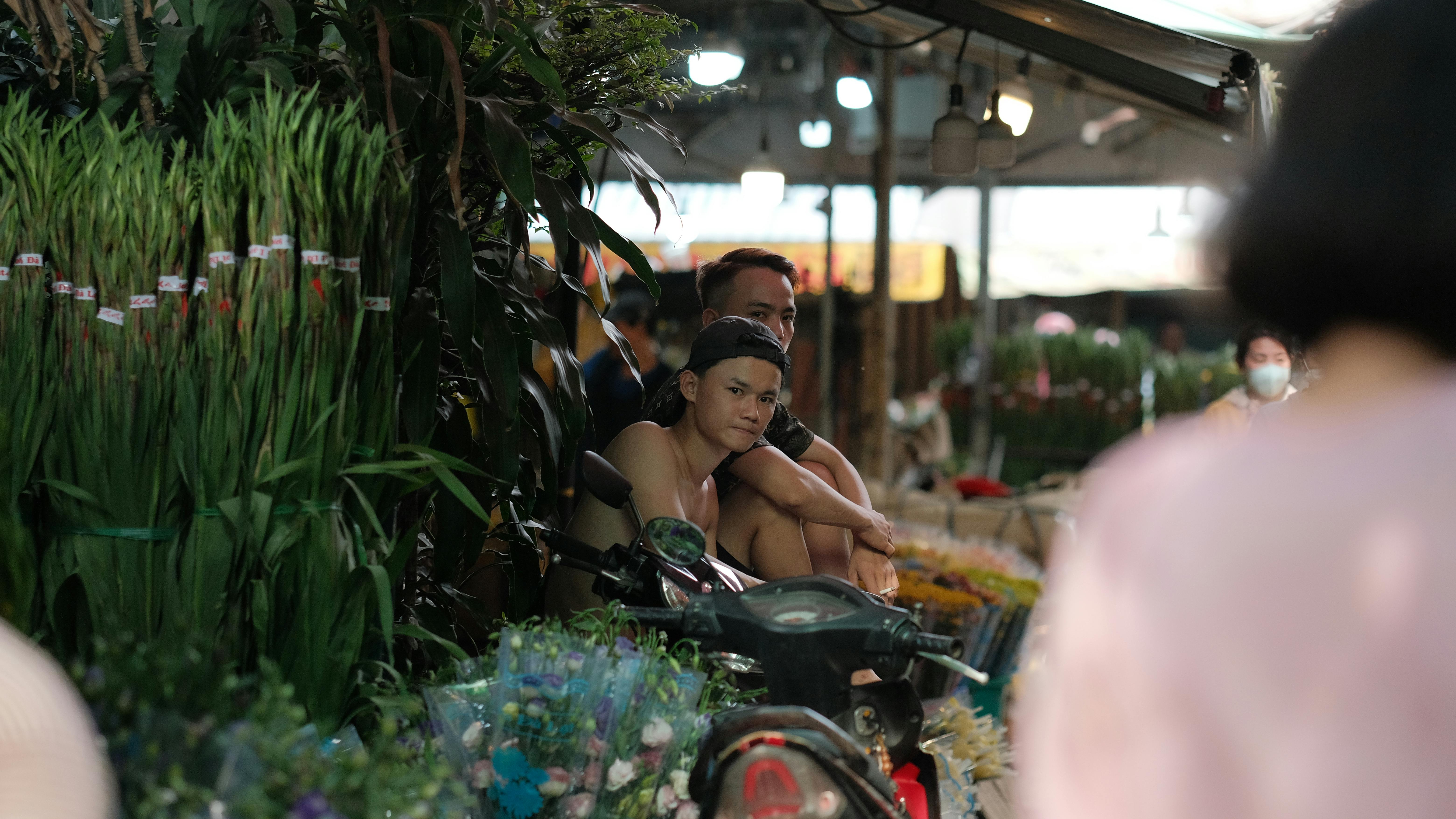 Two people sit near flower stalls in a vibrant market setting.