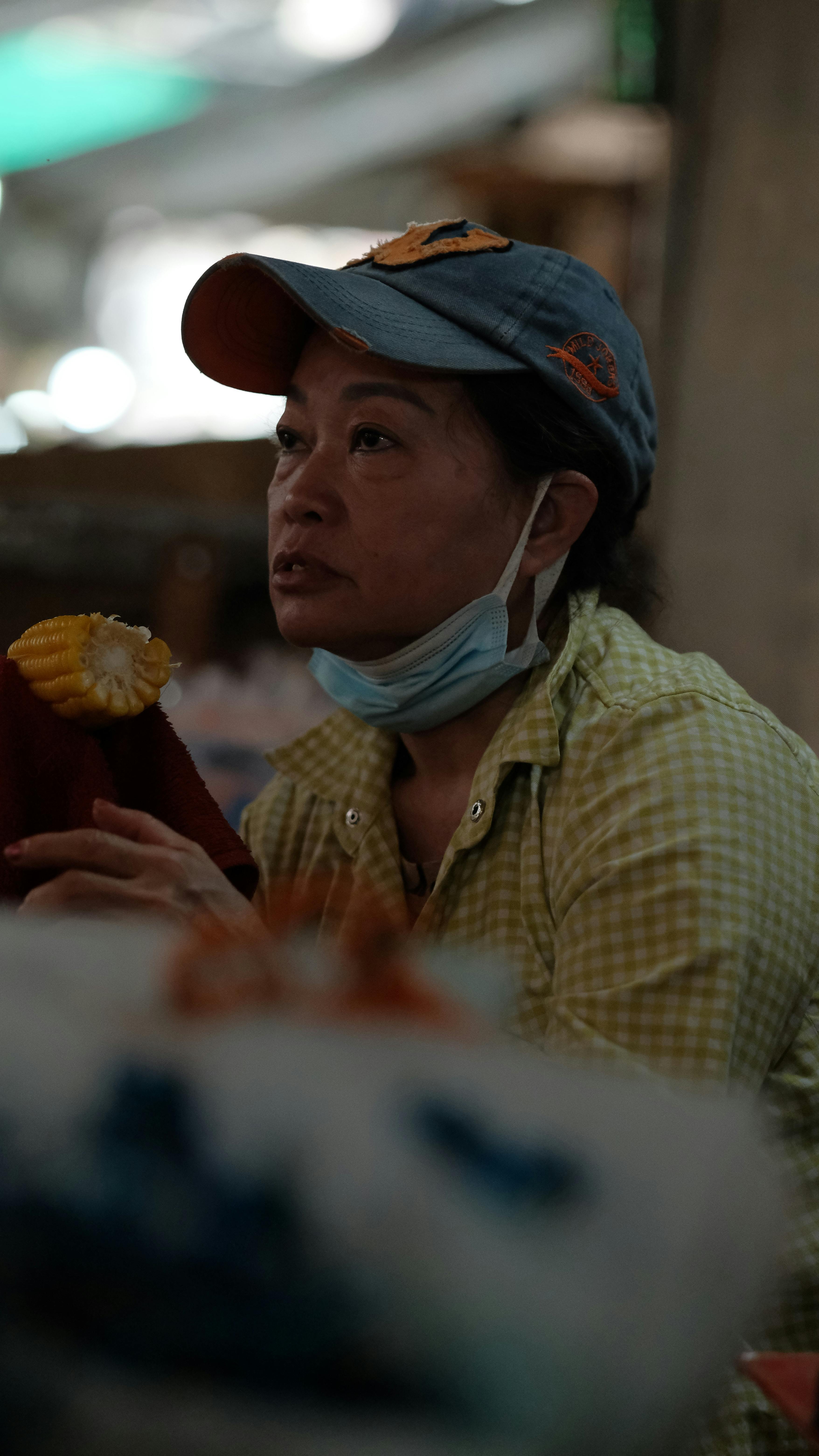A woman in a market setting, wearing a cap and a face mask under her chin, with a piece of corn.