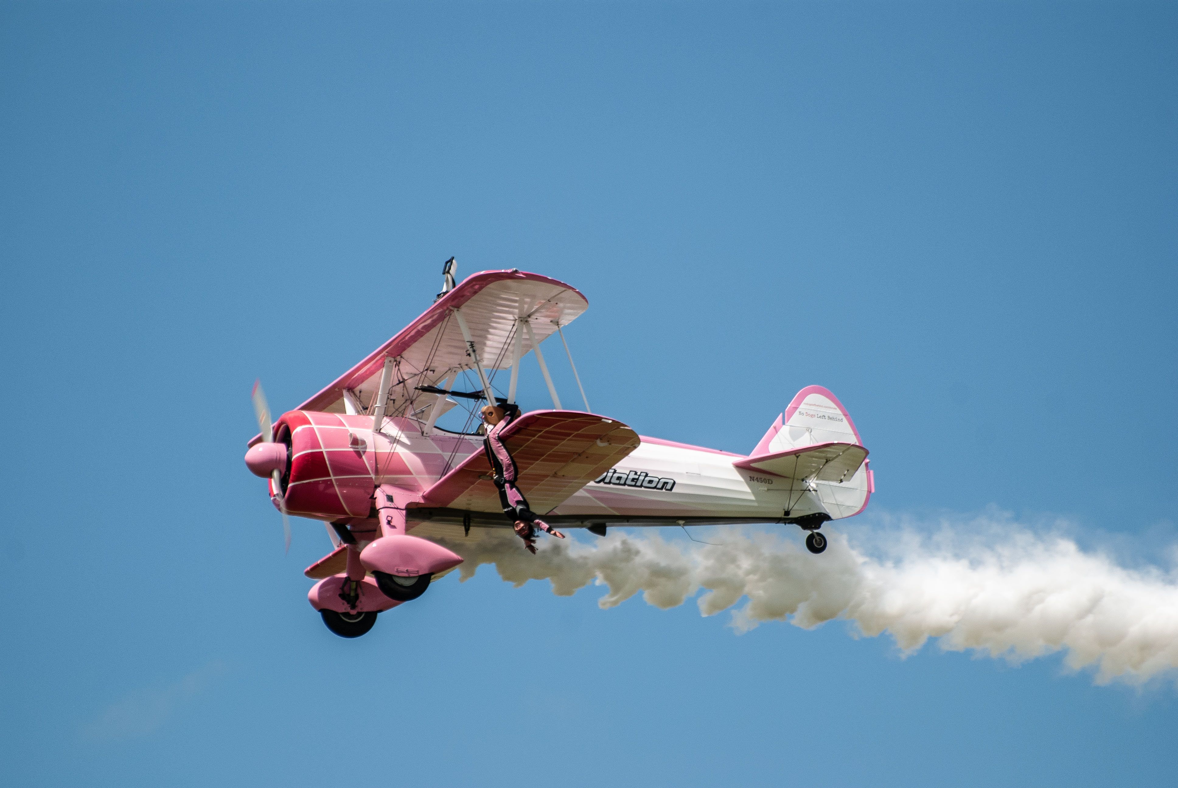 A person performing stunts on the wing of a vintage pink biplane during an airshow.