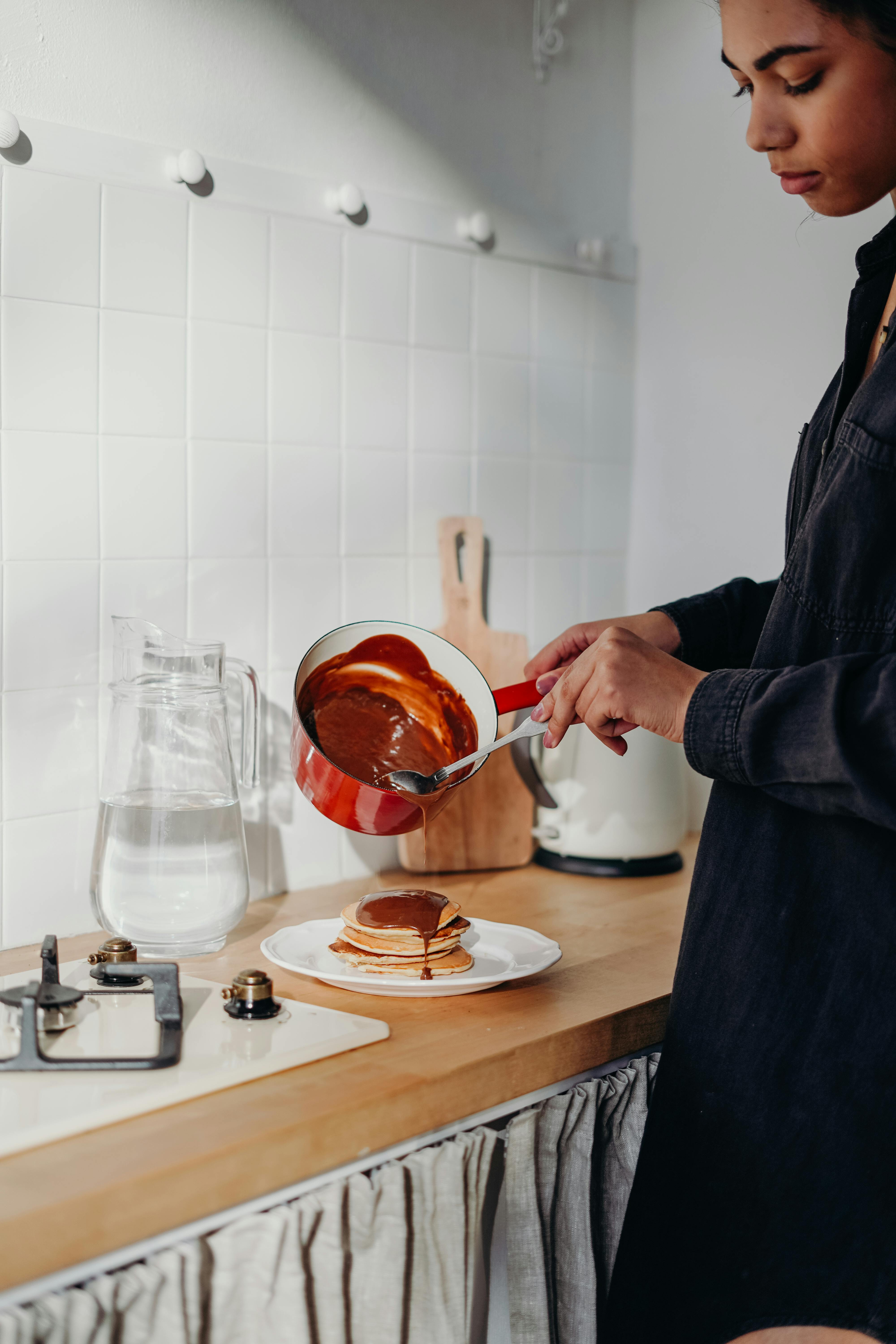 A woman pours chocolate syrup onto pancakes in a cozy kitchen setting, creating a delicious breakfast scene.