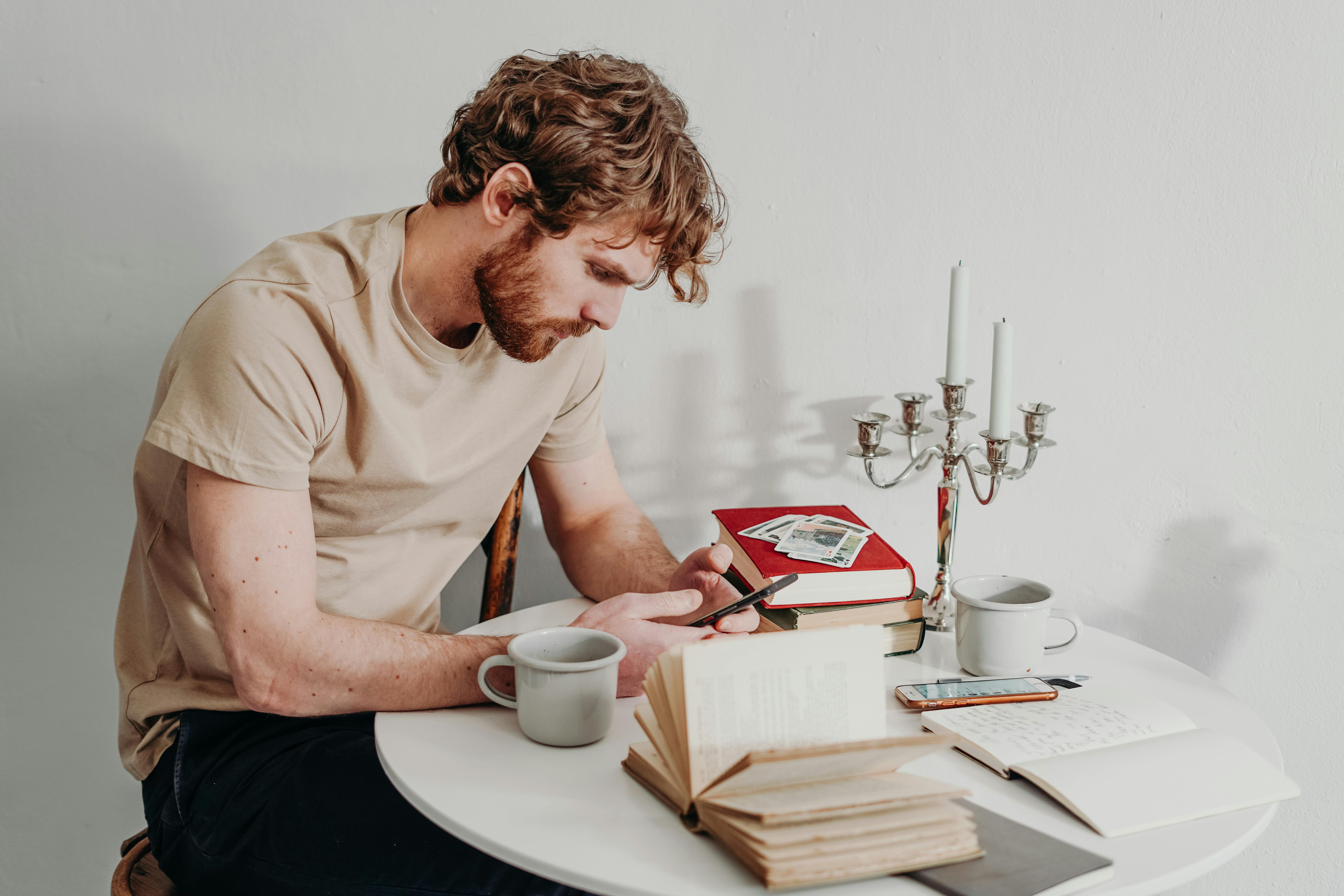 一个男人在室内，一边看书一边用着智能手机