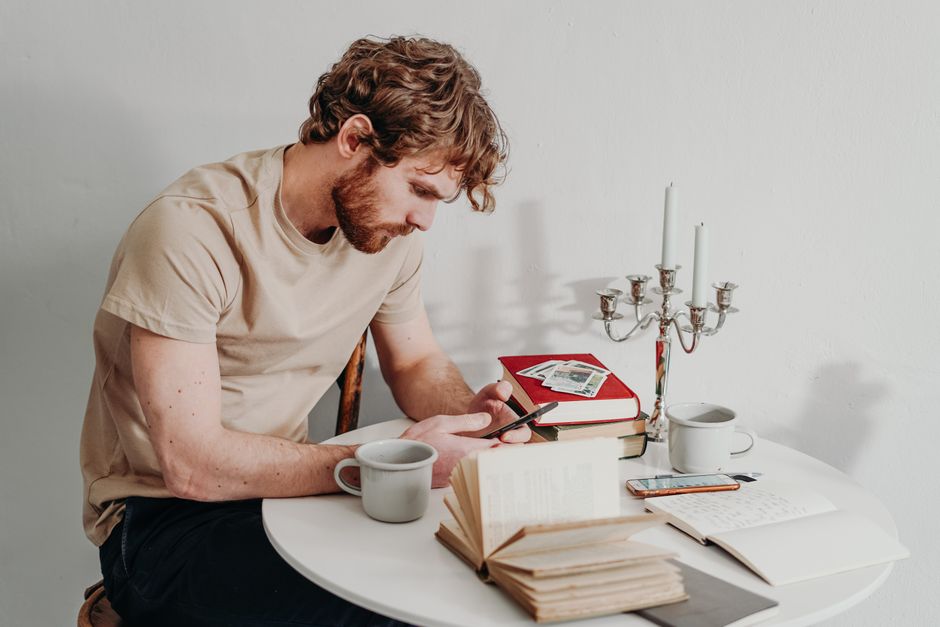A man reads at a table with books and uses his smartphone indoors. A man reads at a table with books and uses his smartphone indoors.