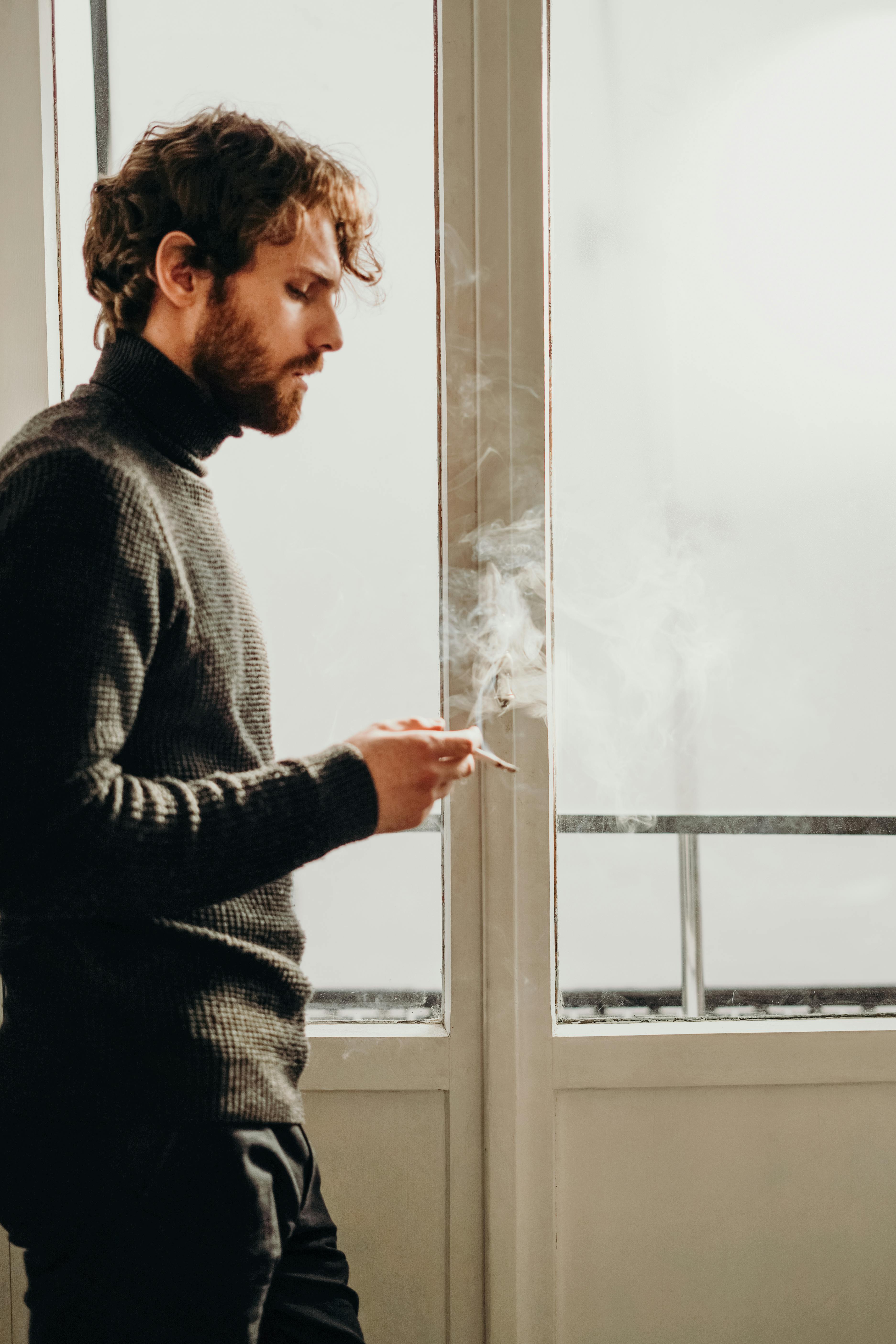 man wearing black sweater standing beside white wooden framed glass window while smoking