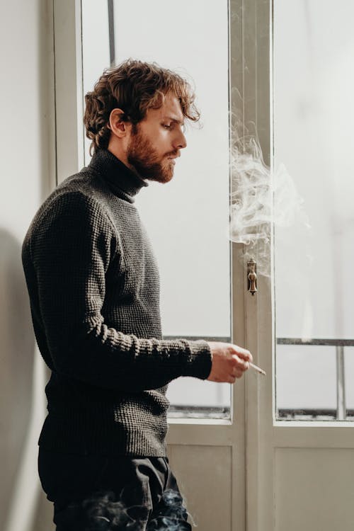Side View Photo of Man in Black Sweater Standing Beside White Wooden Door Smoking
