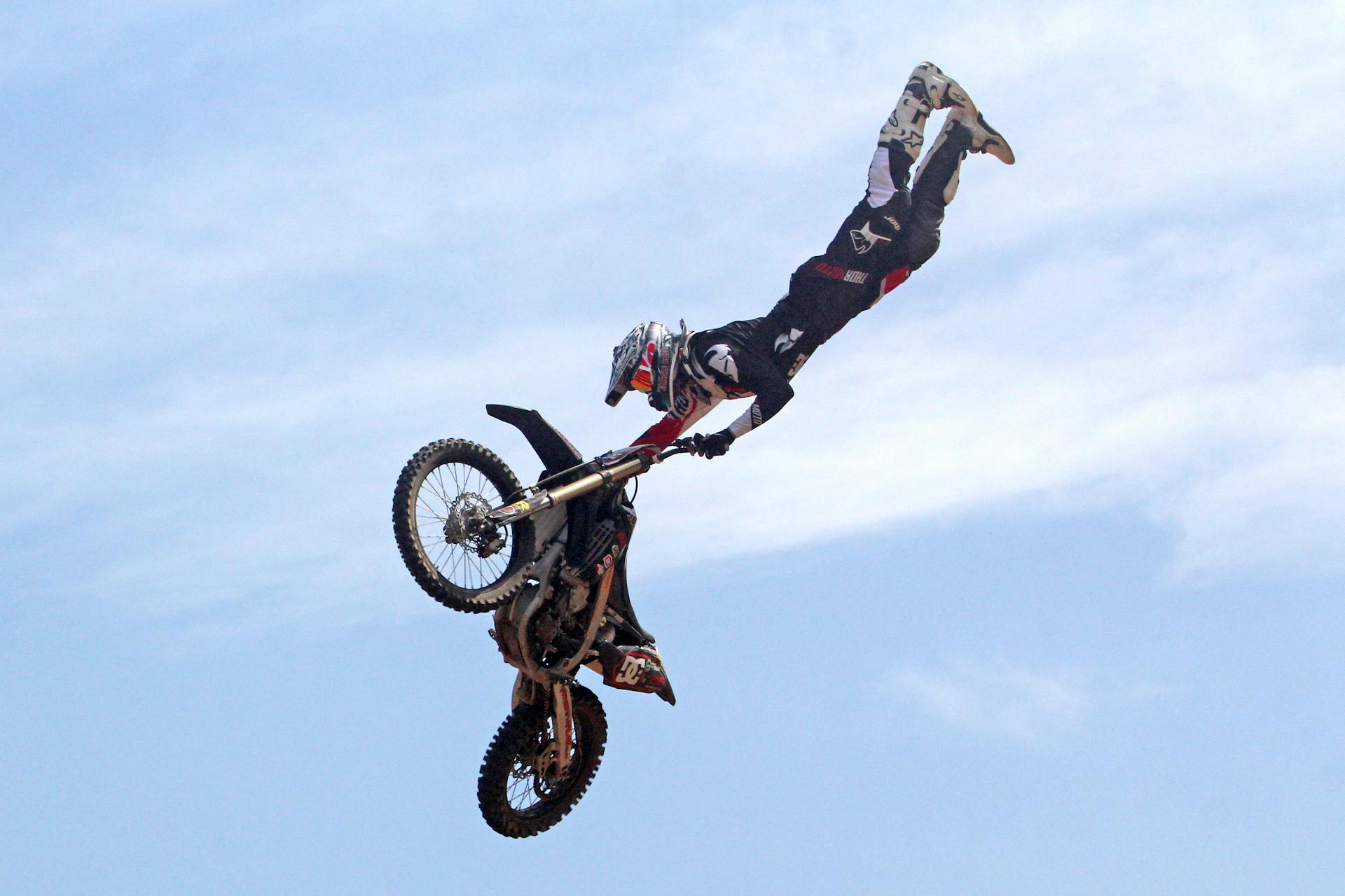 A motocross rider performs a daring freestyle stunt in mid-air against a clear blue sky.