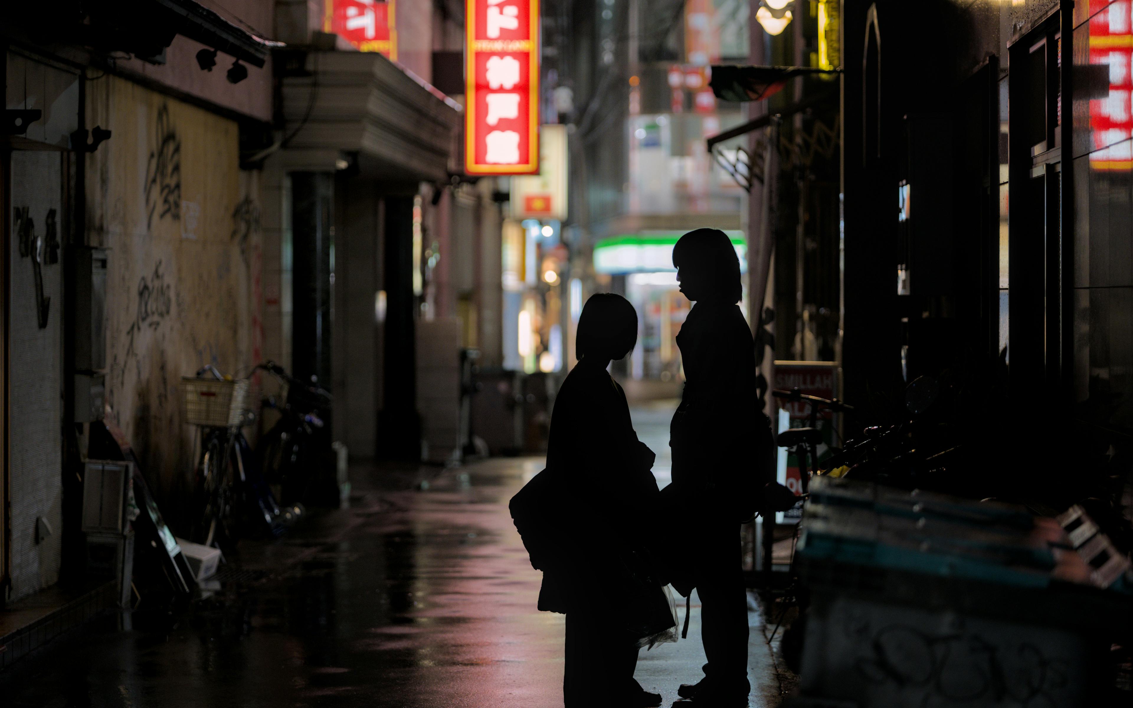 A silhouetted couple stands in a neon-lit alley in Kobe, Japan, at night.