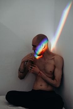 一个秃头纹身男人光着膀子坐在白墙上，被彩虹灯照亮