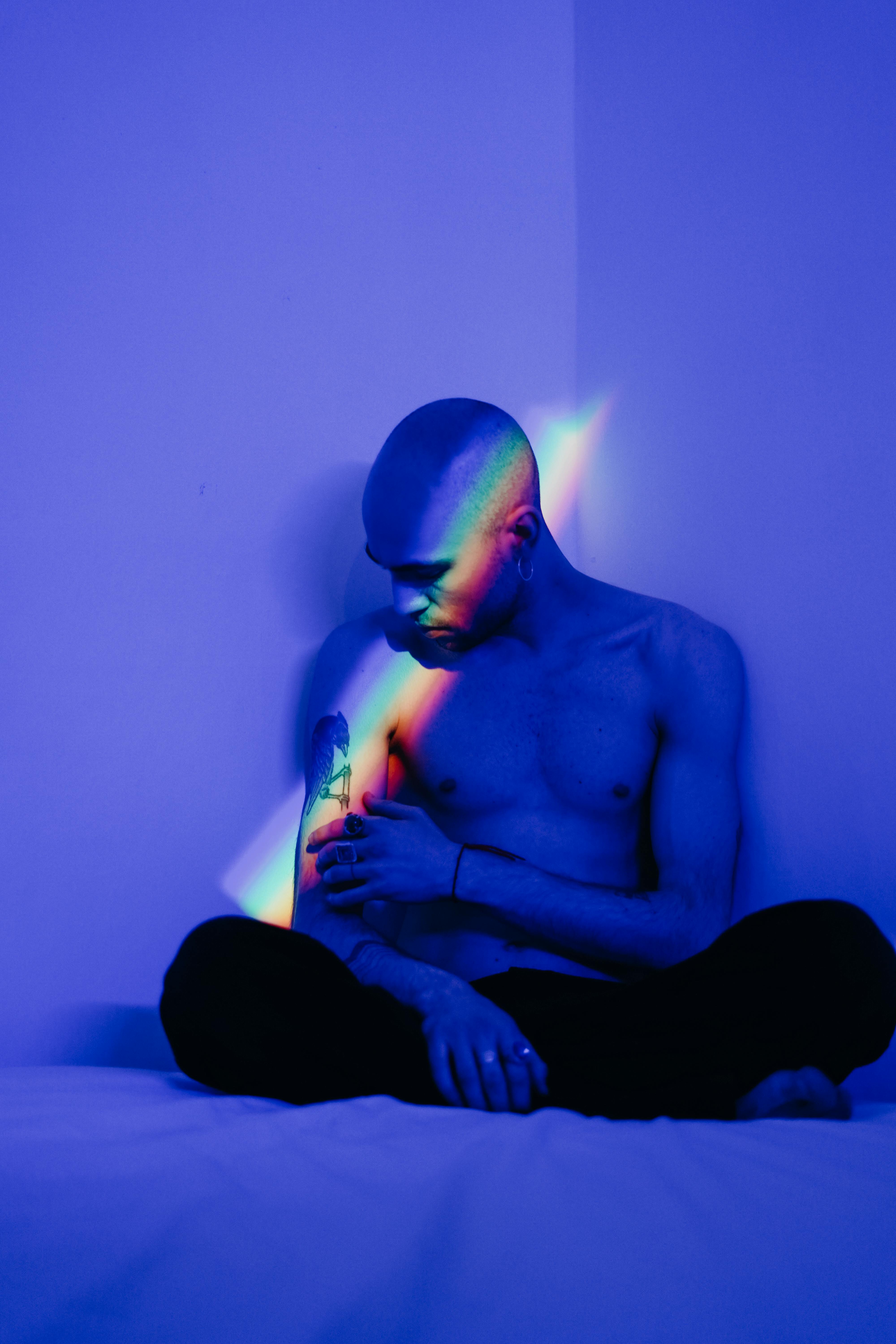 Shirtless tattooed man sitting indoors in blue ambient light with a rainbow reflection crossing his body.