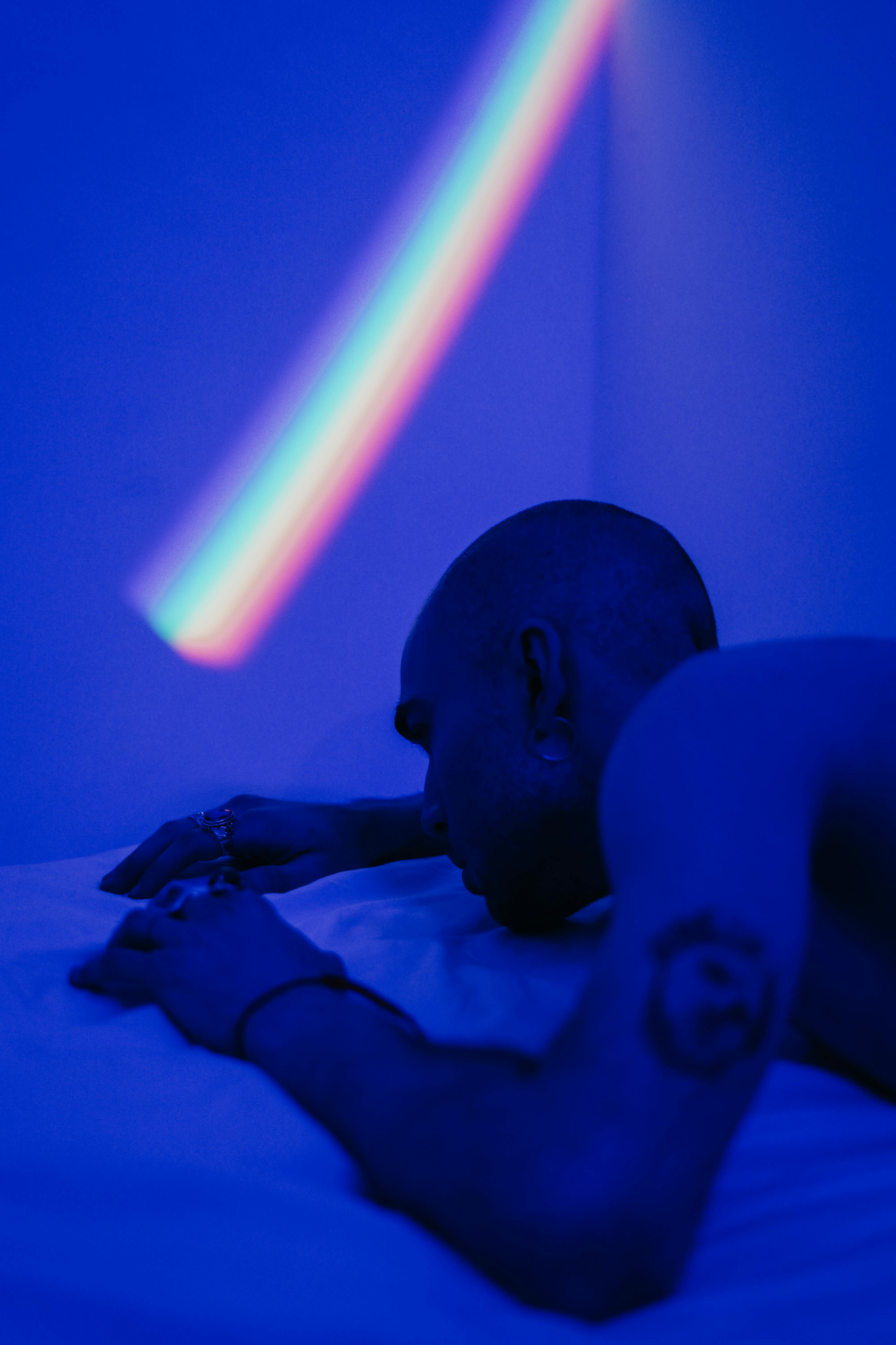 A bald, tattooed man lies shirtless under a rainbow light in a moody blue-lit room.