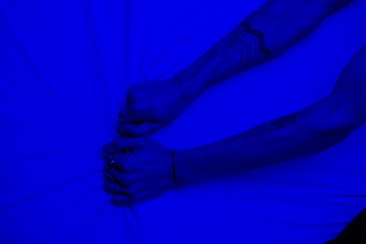 Close-up of tattooed arms pulling fabric under intense blue lighting, symbolizing stress or frustration.