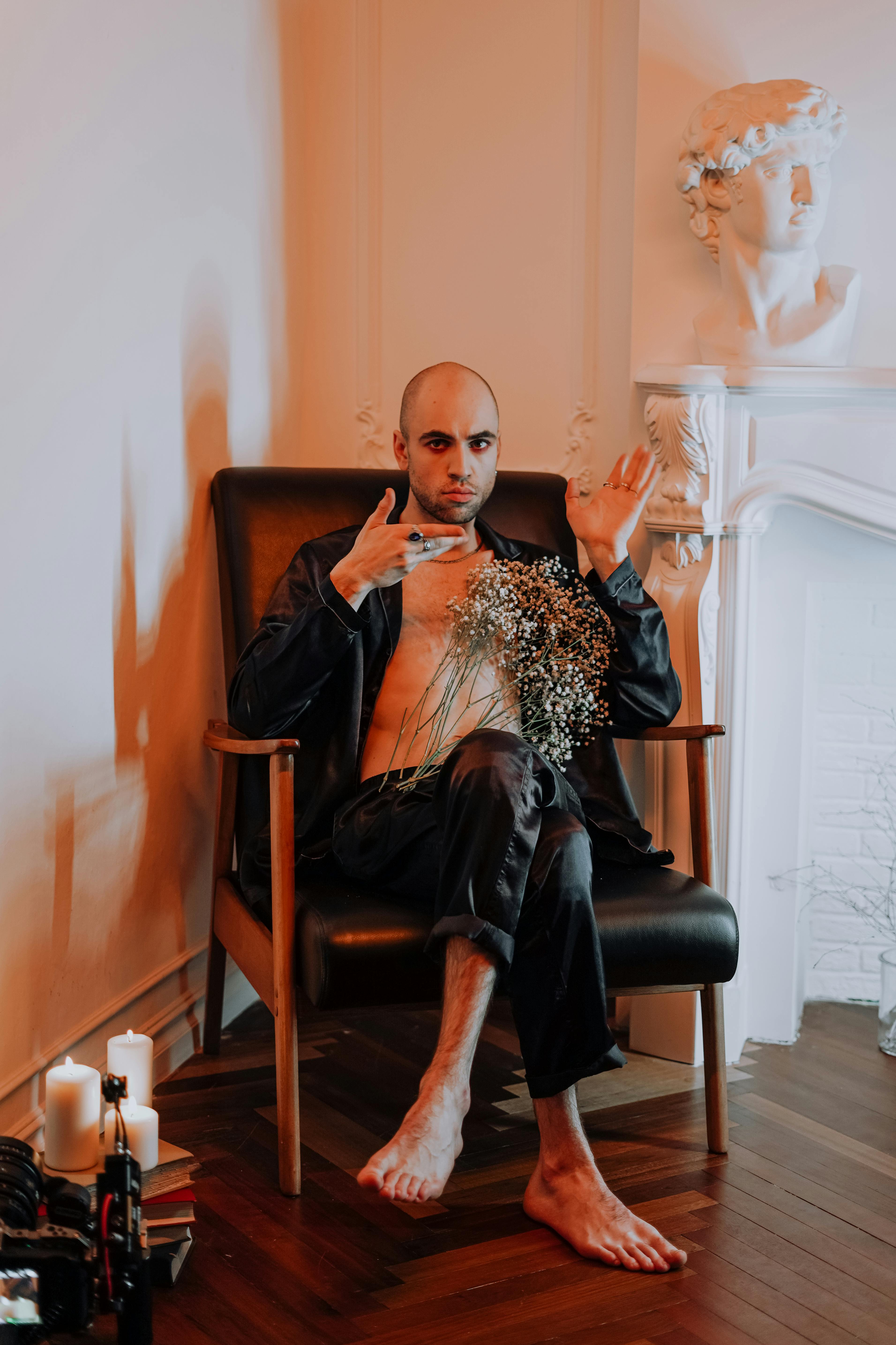 A bald man in black attire poses in an armchair, holding flowers, in a stylish indoor setting.