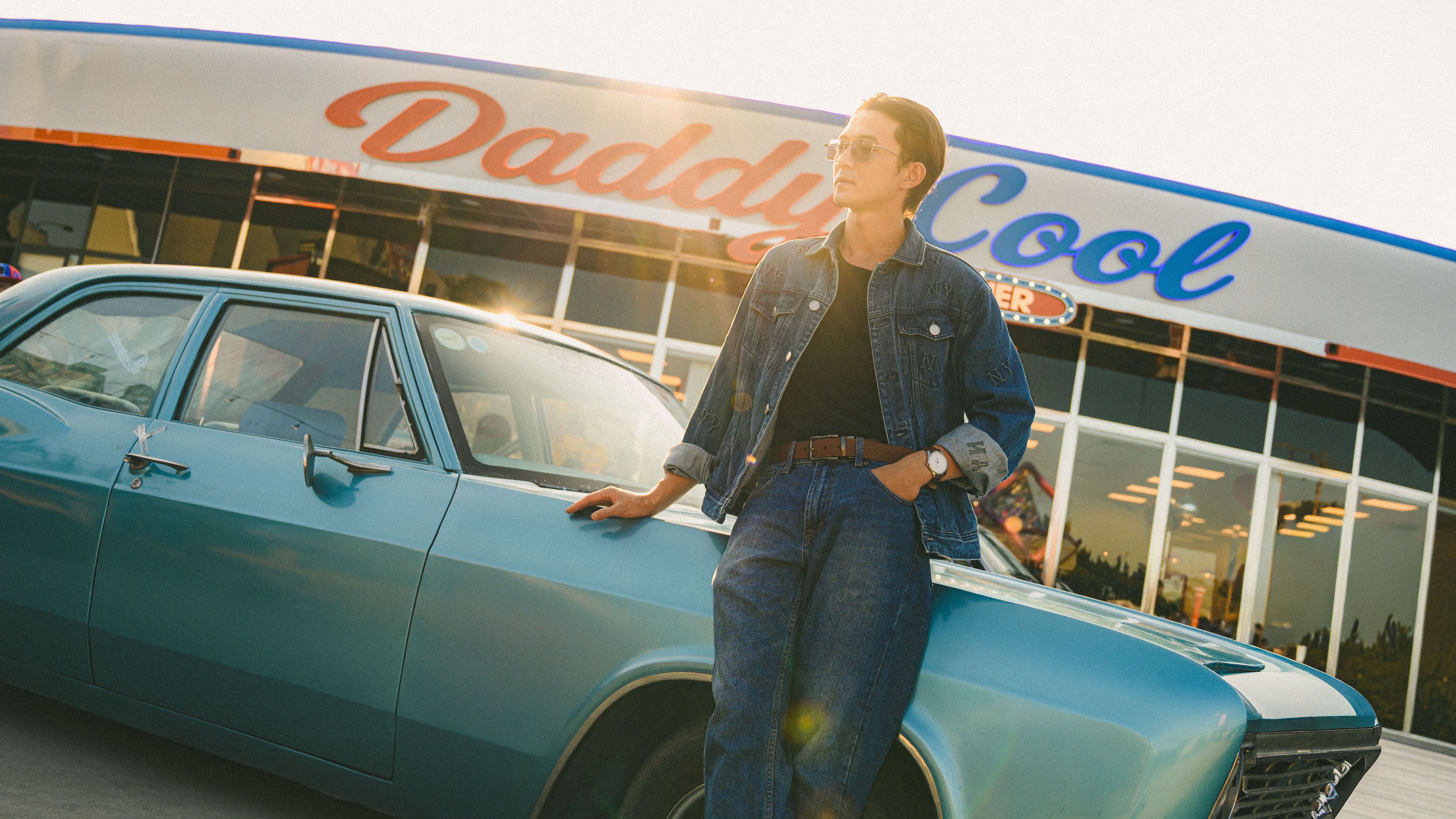 Man in denim outfit leaning on classic blue car at retro diner.