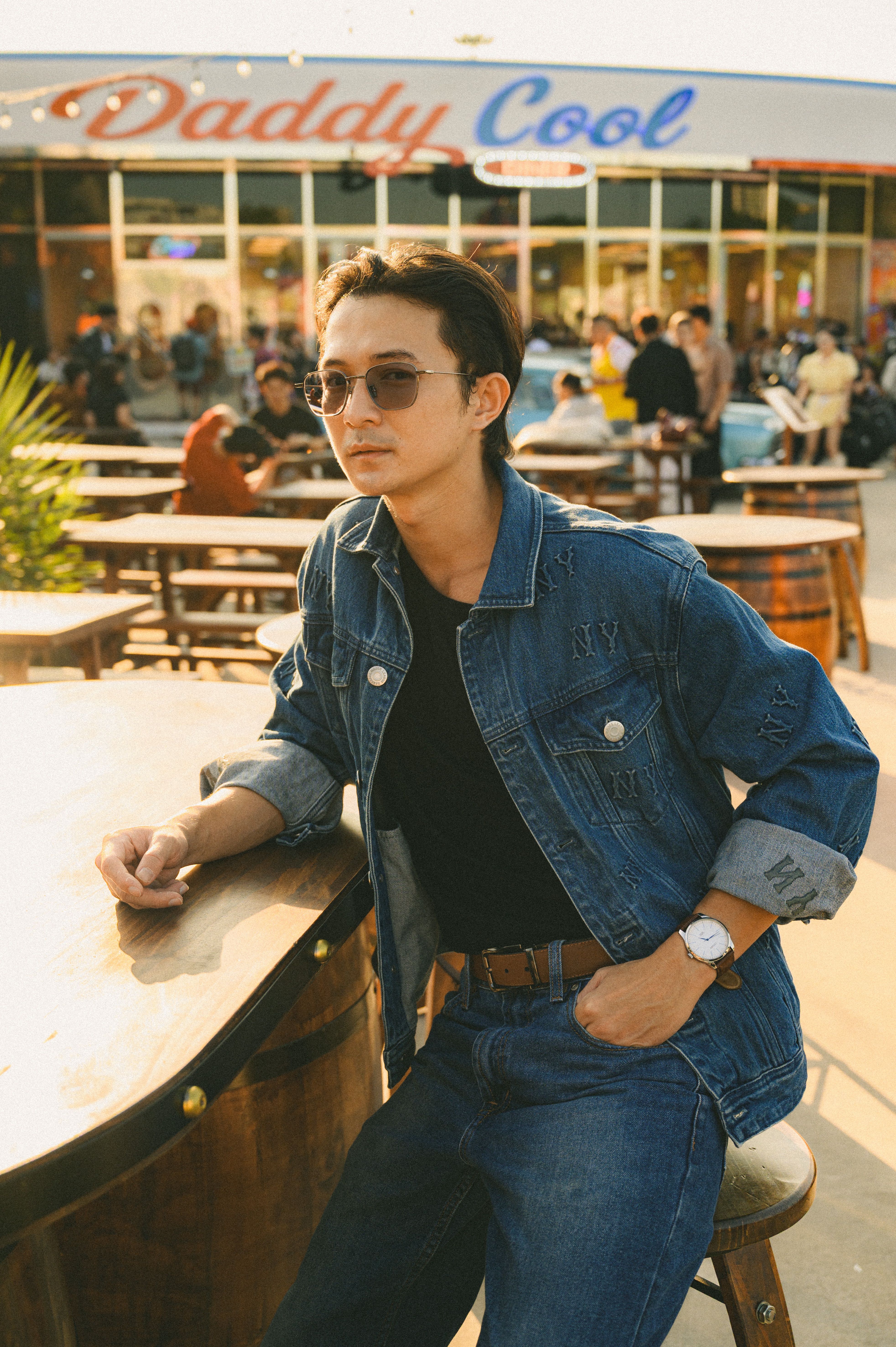 Stylish young man in denim jacket and sunglasses at a sunny outdoor cafe.