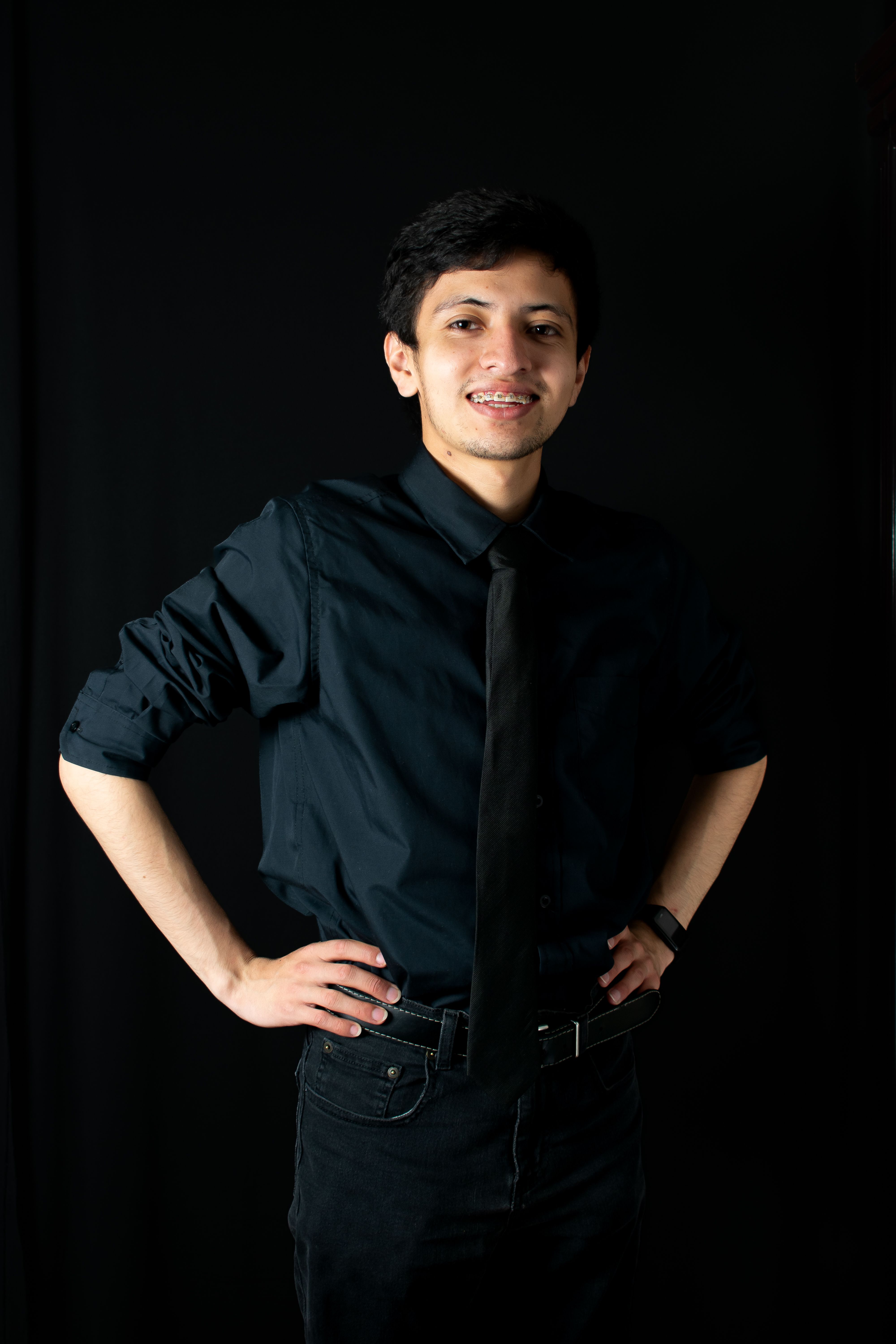 Portrait of a young man in black shirt and tie against a dark backdrop, smiling confidently.
