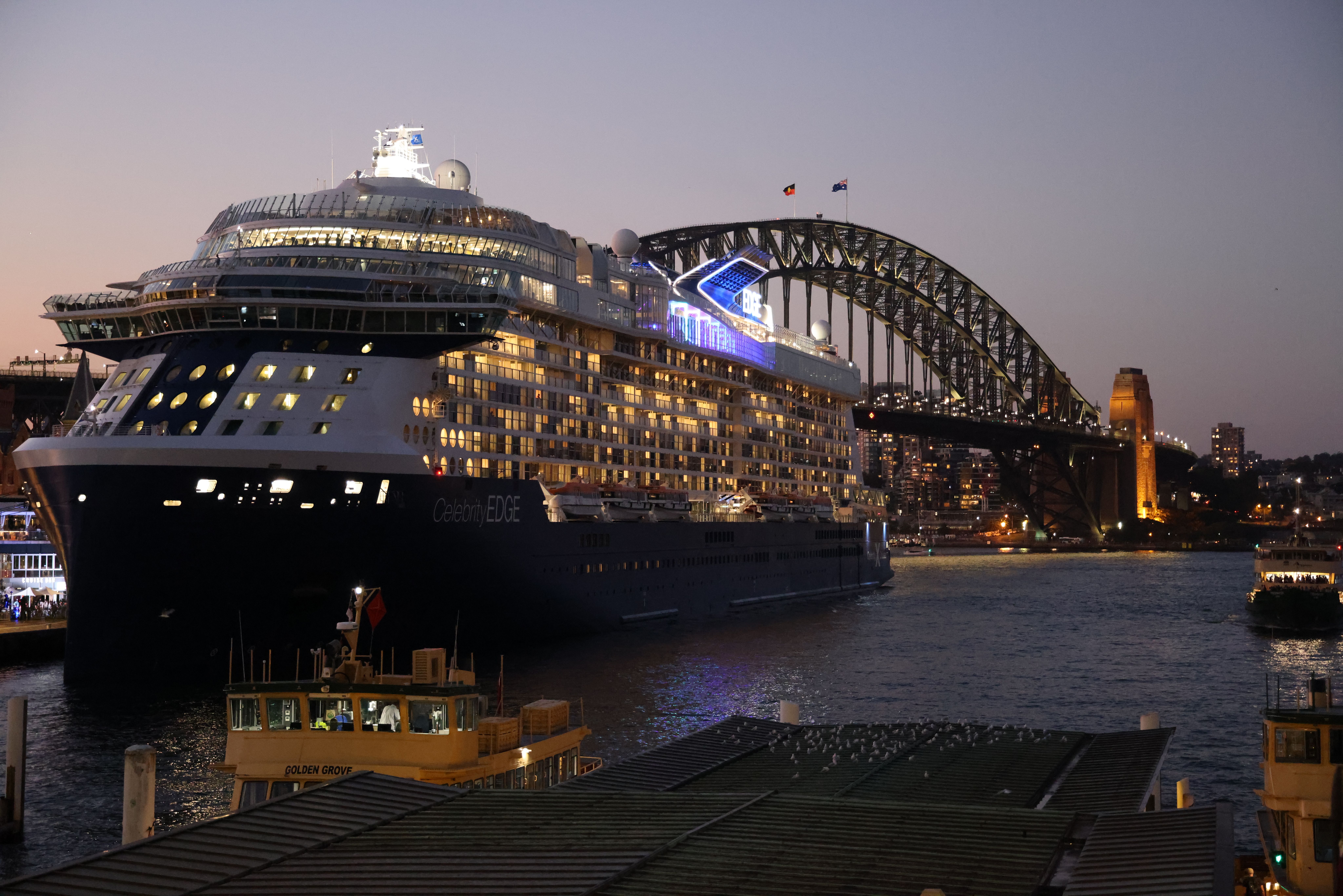 A large cruise ship, Celebrity Edge, sailing past the iconic Sydney Harbour Bridge at dusk.